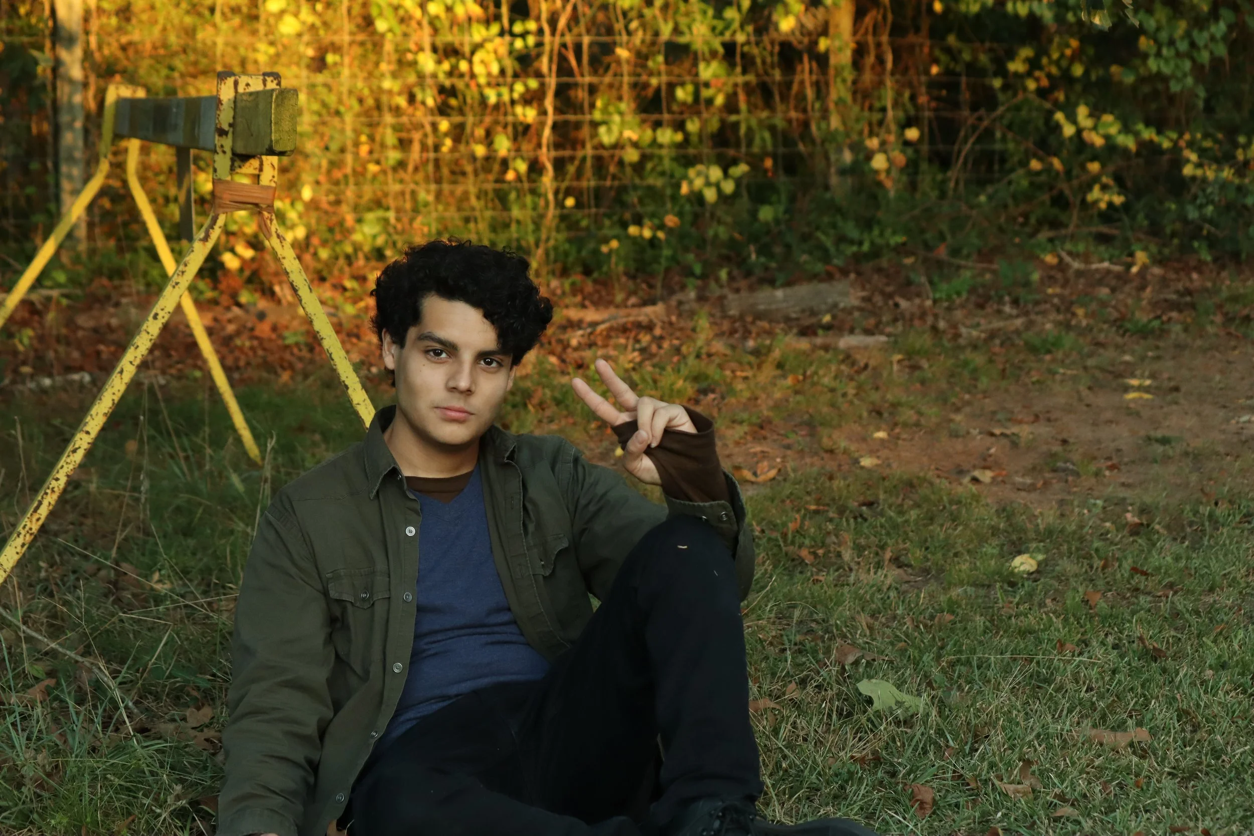 A young man sitting outdoors on grass, making a peace sign with his right hand, next to an old yellow playground slide, with trees and bushes in the background during daylight.
