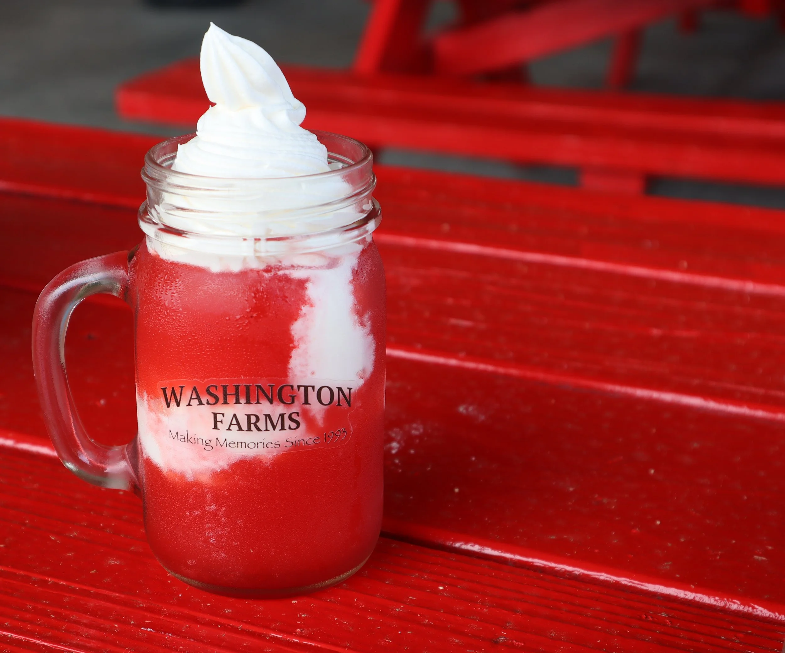A glass jar with foam on top of whipped cream, placed on a red wooden surface with a red bench in the background.