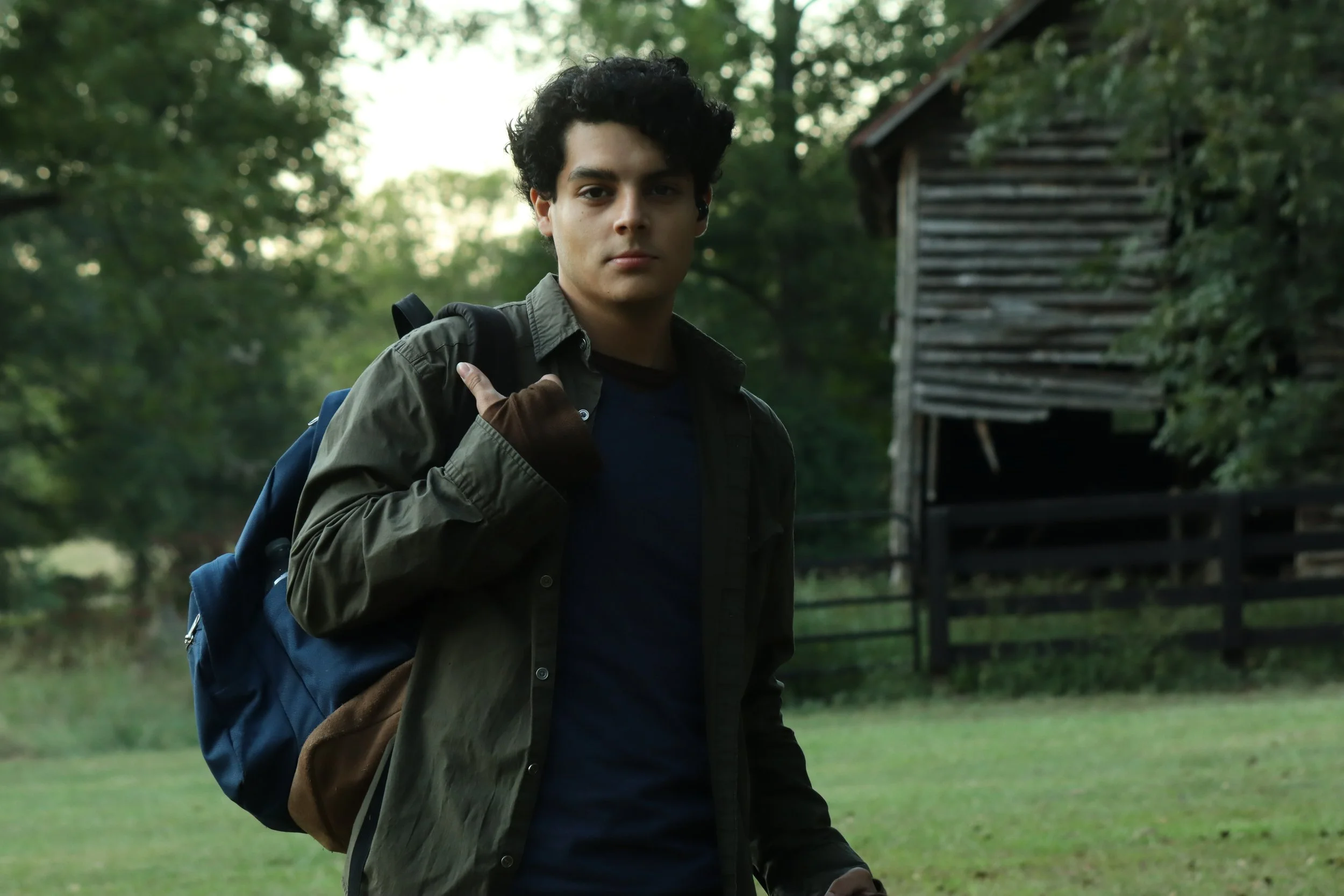 A young man with curly dark hair carrying a backpack over his shoulder, walking outdoors in a green, wooded area near a rustic wooden building.