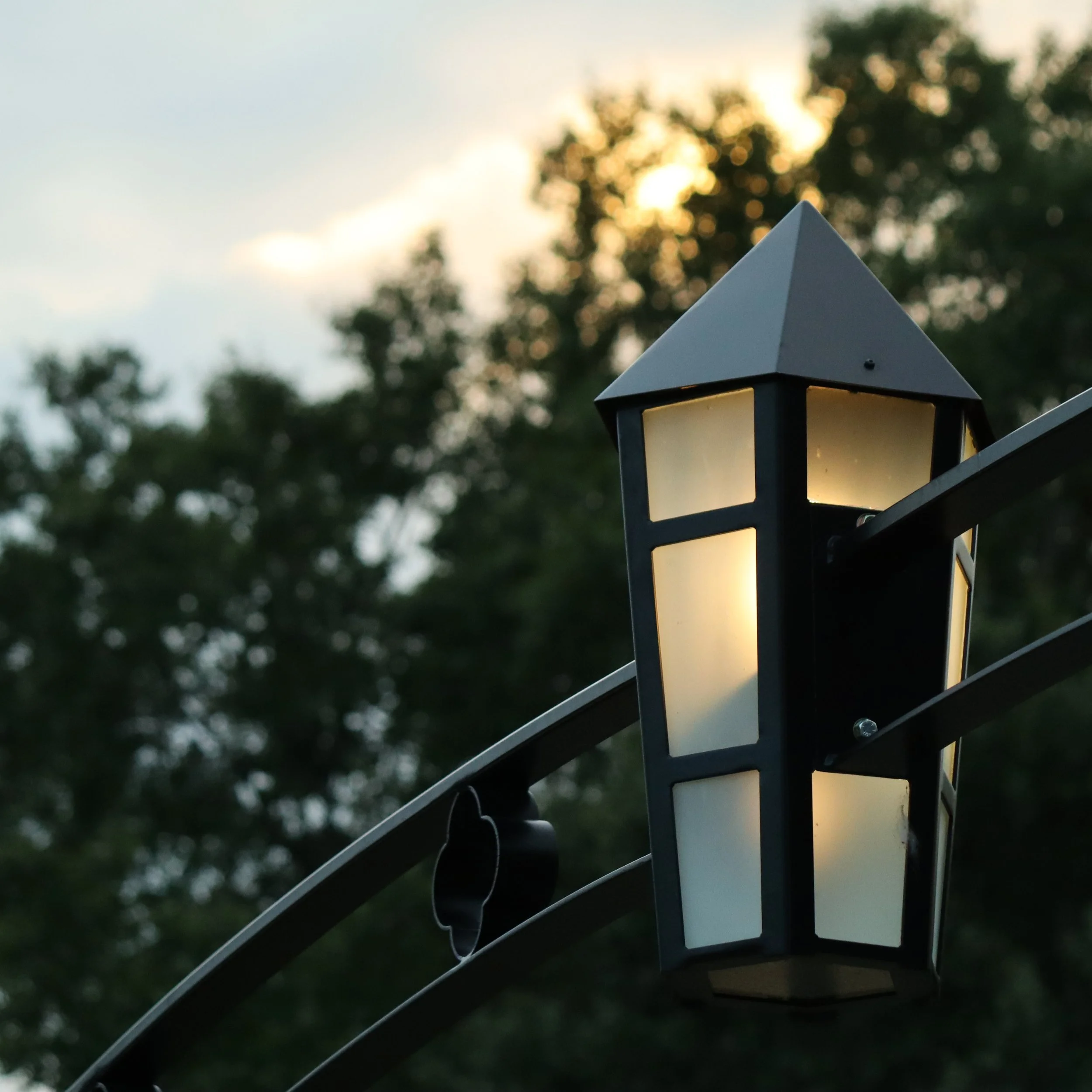 Close-up of a black outdoor lantern with a white frosted glass pane, mounted on a dark metal structure, with a blurred background of trees and a cloudy sky during sunset.