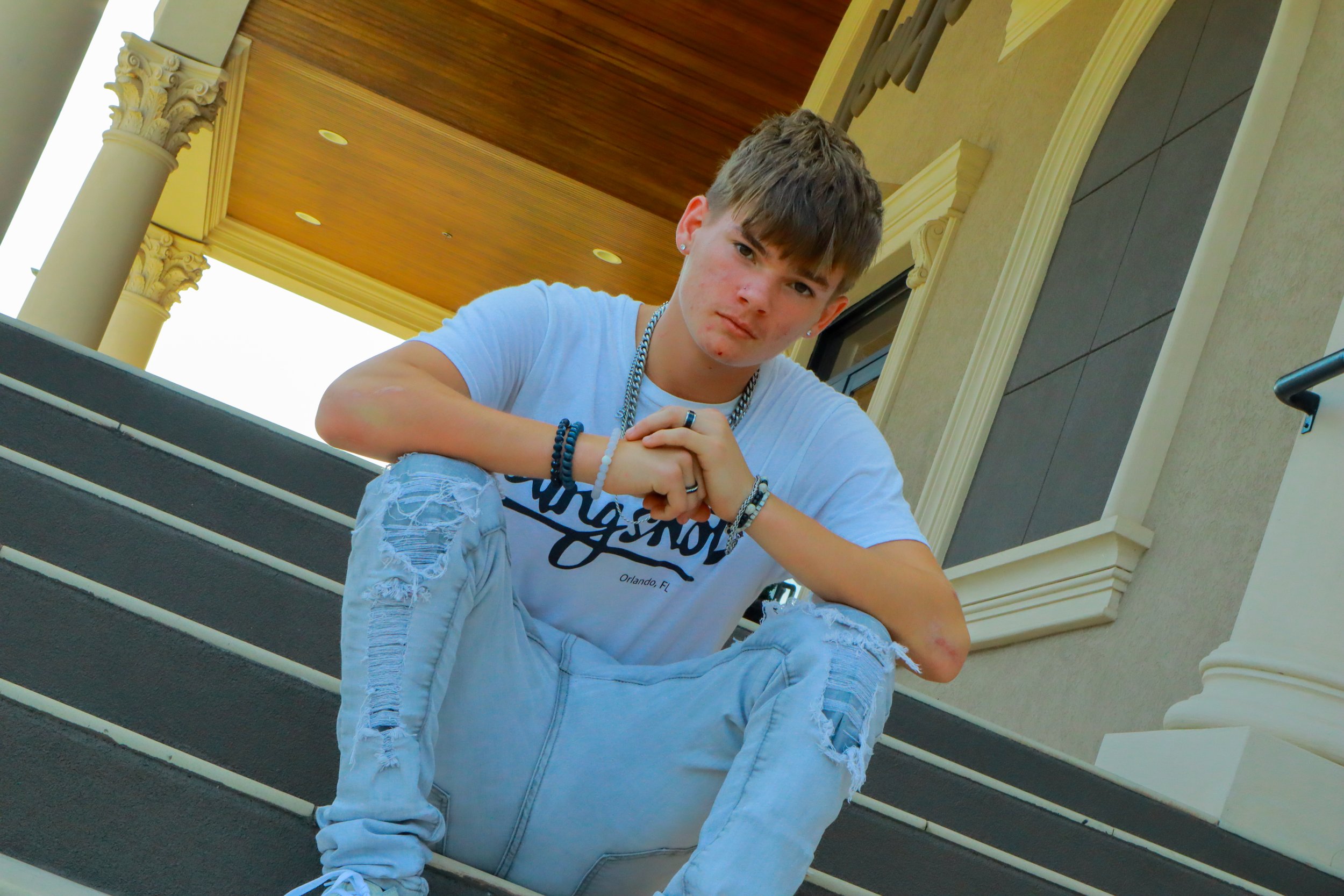 A teenage boy with short brown hair and earrings sitting on outdoor stairs, wearing a light blue T-shirt, ripped jeans, and several bracelets, with a serious expression on his face.