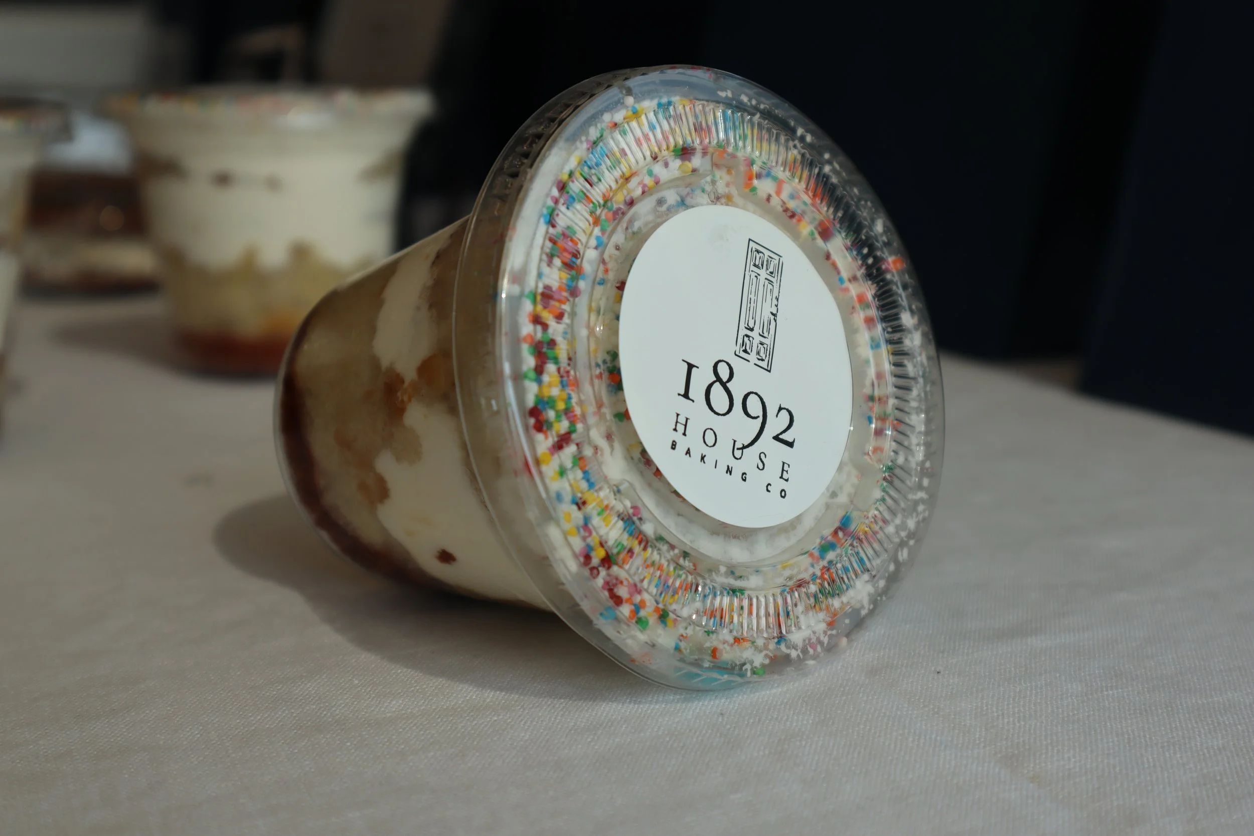 Close-up of a jar with colorful sprinkles on top, labeled '1892 House Baking Co.', placed on a white surface with other dessert jars blurred in the background.