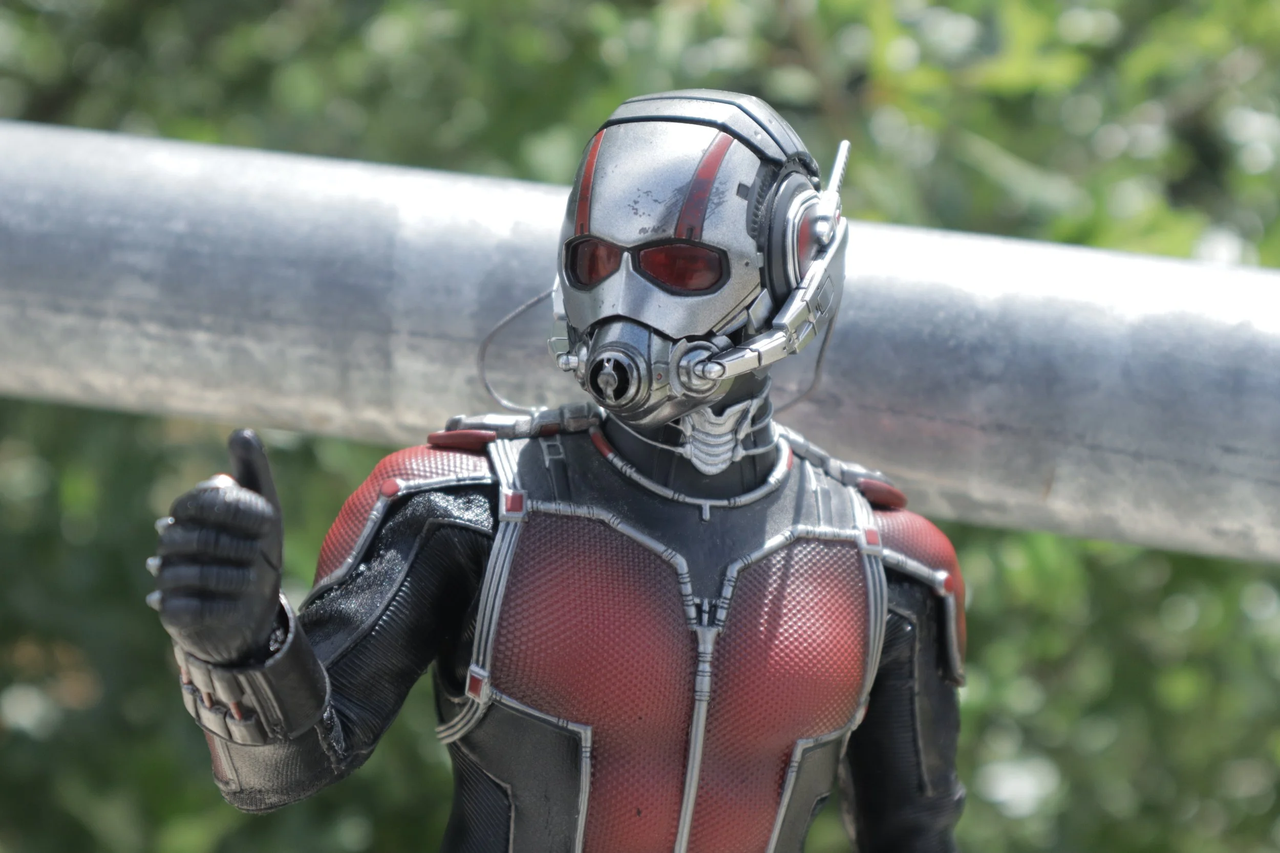 Close-up of Ant-Man from Marvel in his red and black suit with a detailed helmet, pointing his finger forward outdoors with a blurred green background.