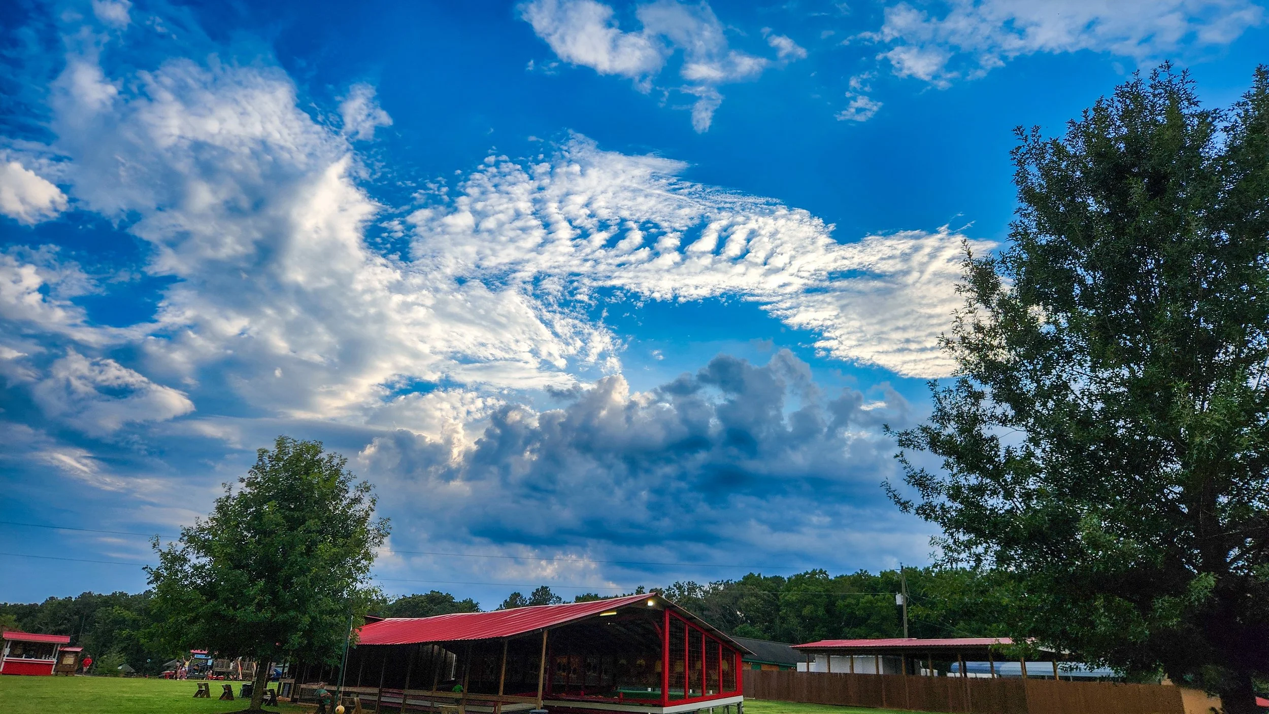 A partly cloudy sky with blue and white clouds above a rural scene with trees, a red-roofed building, and a covered pavilion area.
