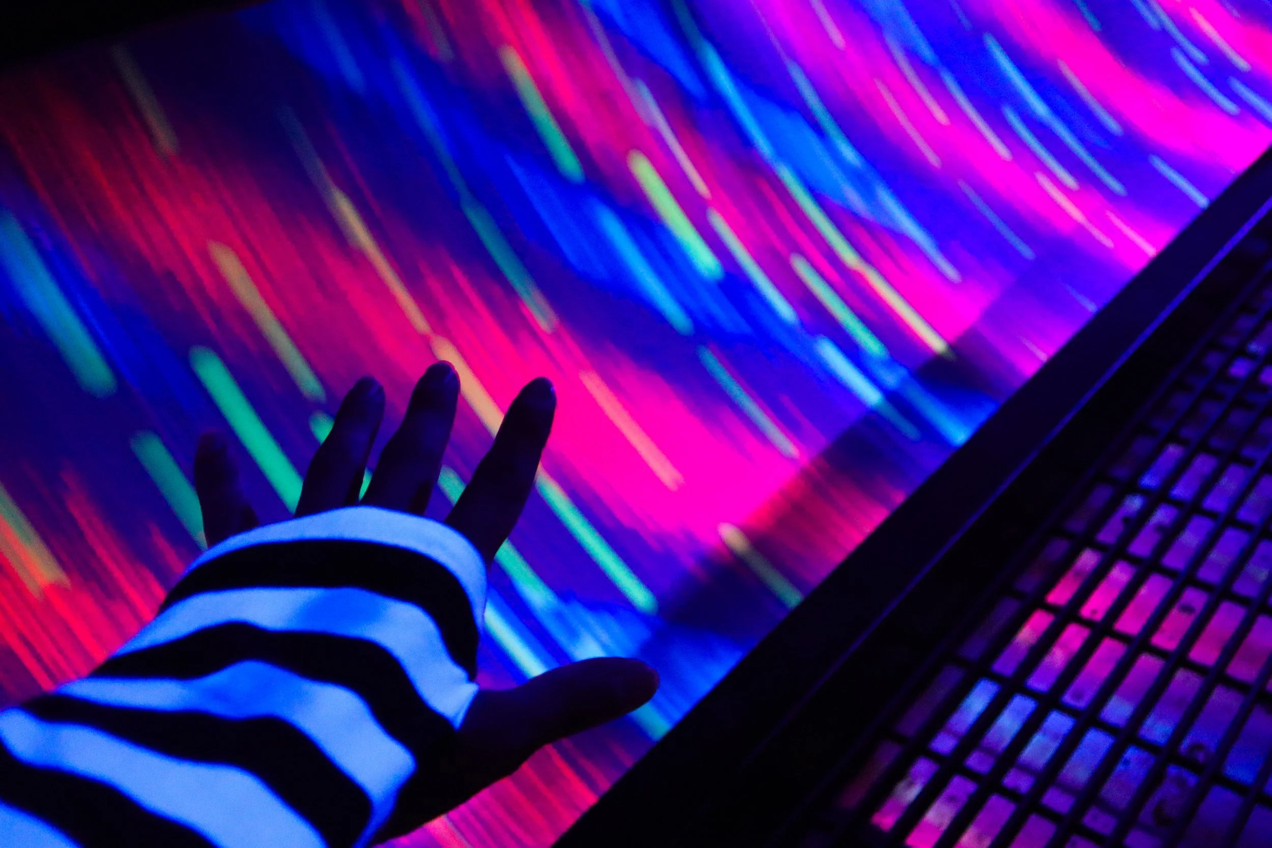 Hand with a striped sleeve reaching out towards a colorful, neon-lit surface with diagonal streaks of pink, blue, purple, and yellow.