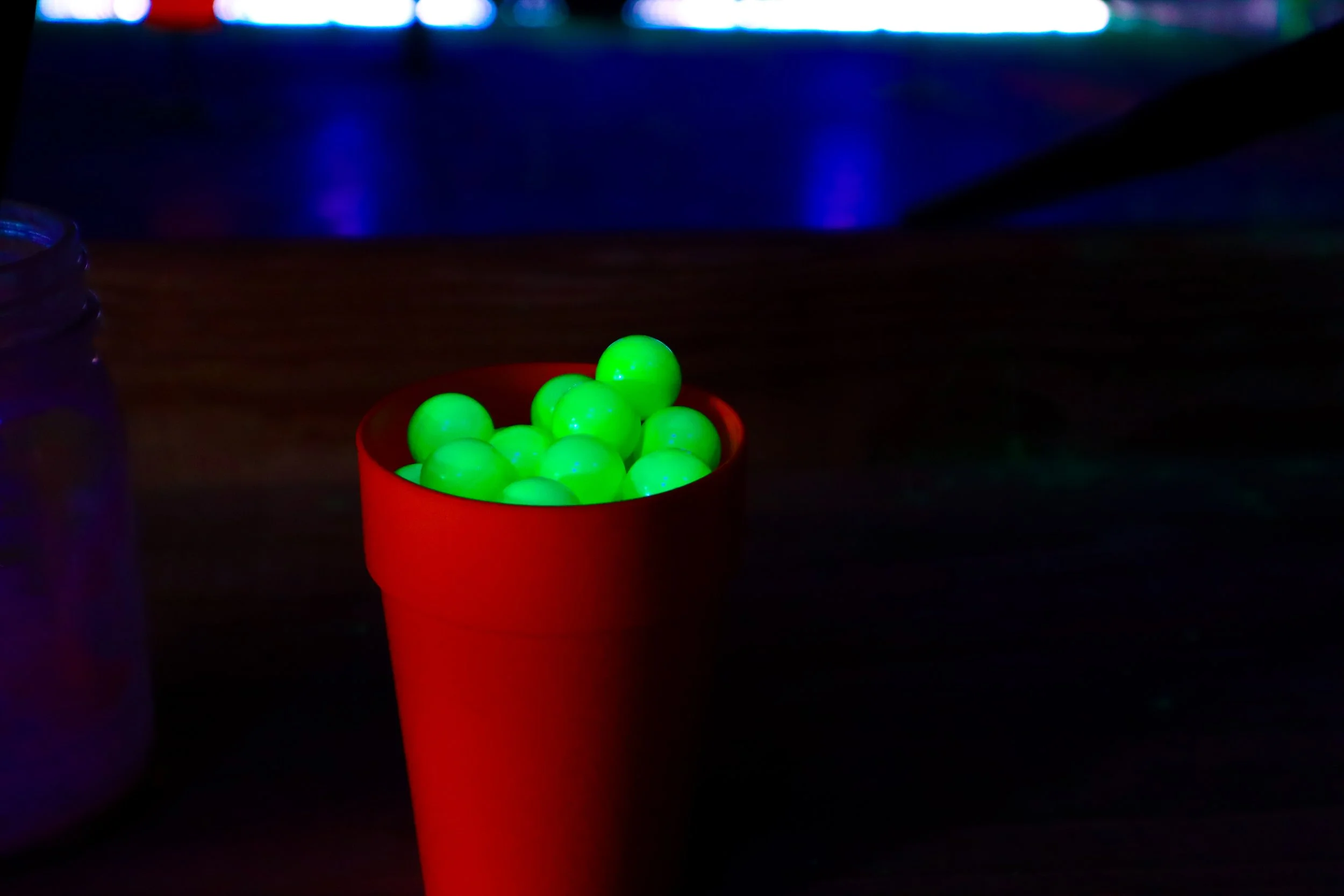 A red cup filled with glow-in-the-dark green balls in a dark environment with a blurred background of colorful lights.