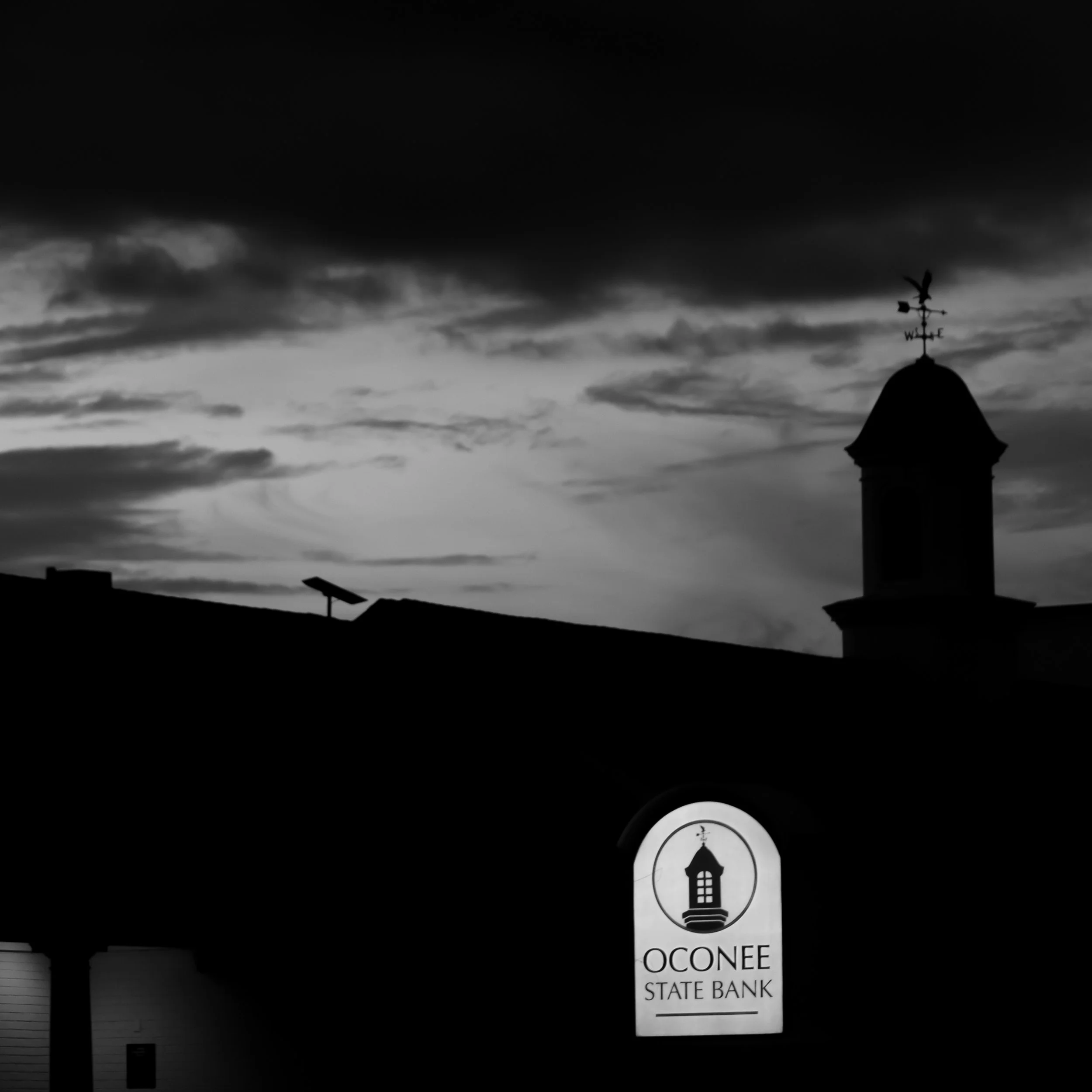A weather vane with a rooster on top, located on a building. A vertical sign reads 'OCONEE STATE BANK,' with a logo of a tower and the bank's name below. The sky is cloudy and dark.