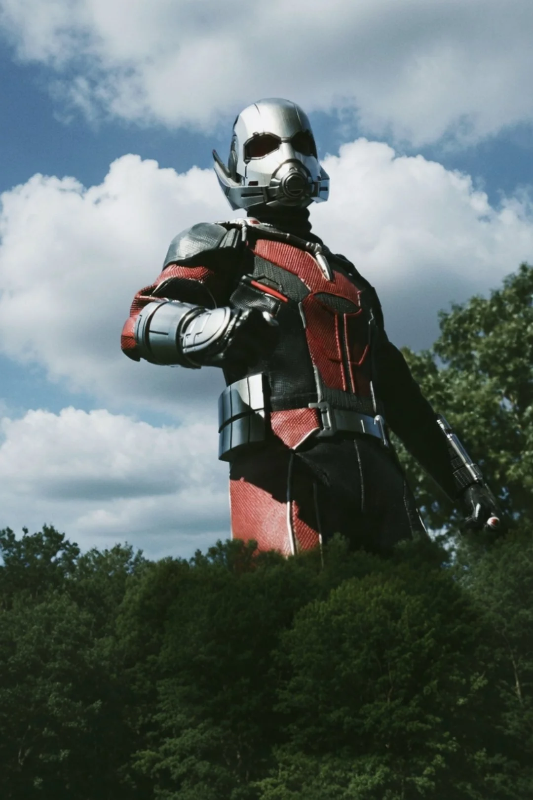 A person dressed as Ant-Man standing outdoors with a blue sky, white clouds, and green trees in the background.