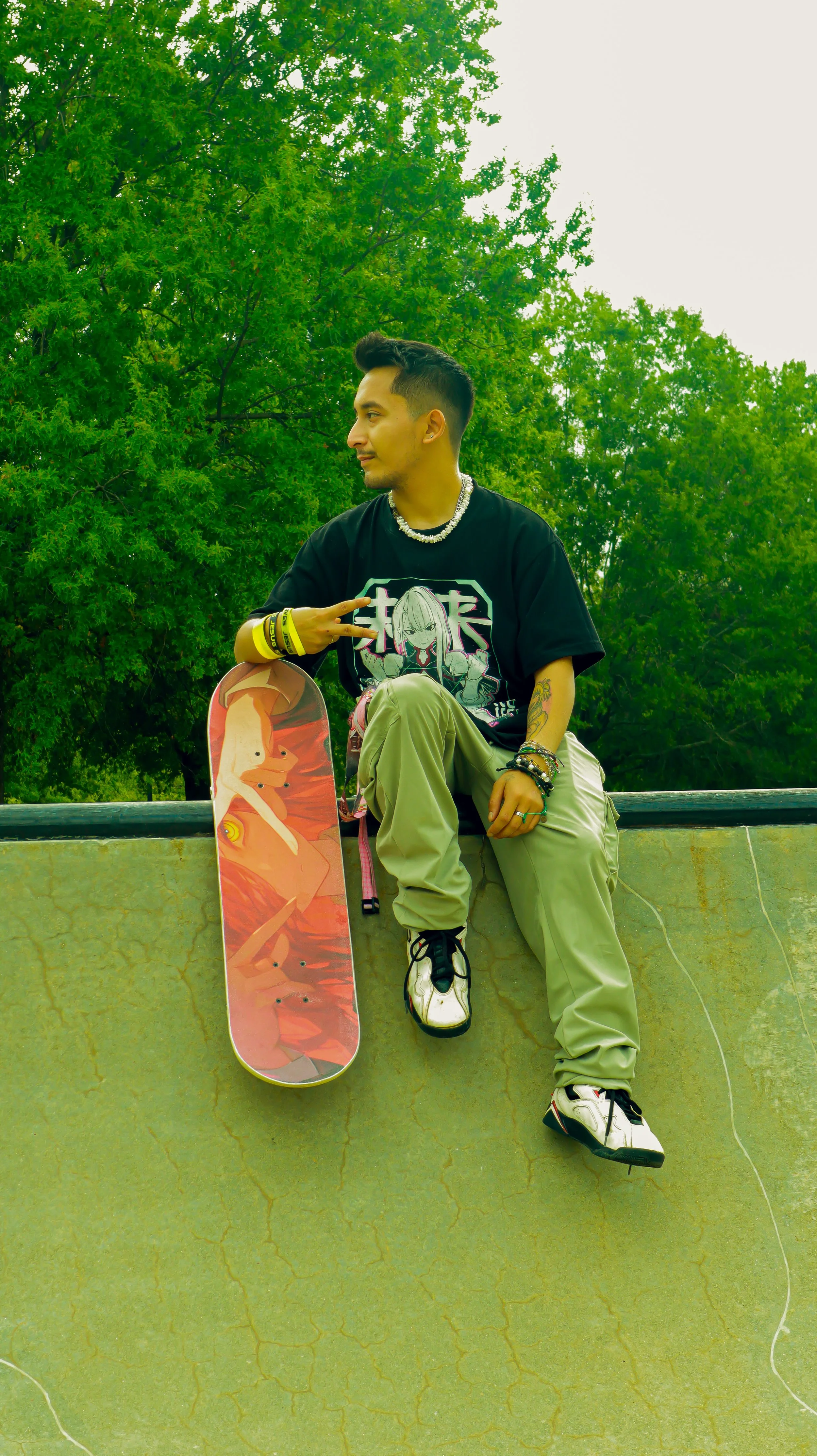 A young man with dark hair, wearing a black t-shirt with anime art, light green pants, and sneakers, sitting on a skatepark ledge with a skateboard. He is making a peace sign with his right hand and looking to his left. The background features lush g