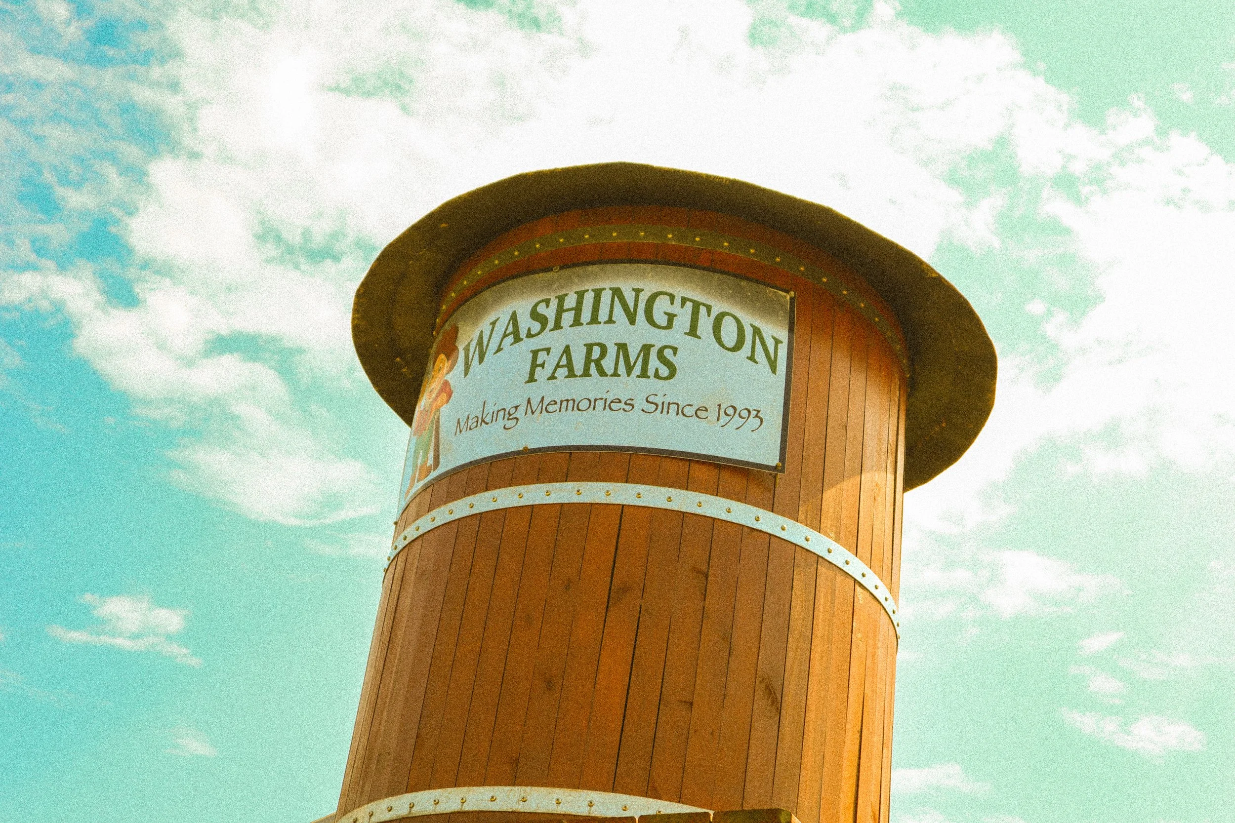 Tall wooden water tower with a rounded top, featuring a sign that reads 'Washington Farms Making Memories Since 1993,' set against a partly cloudy sky.