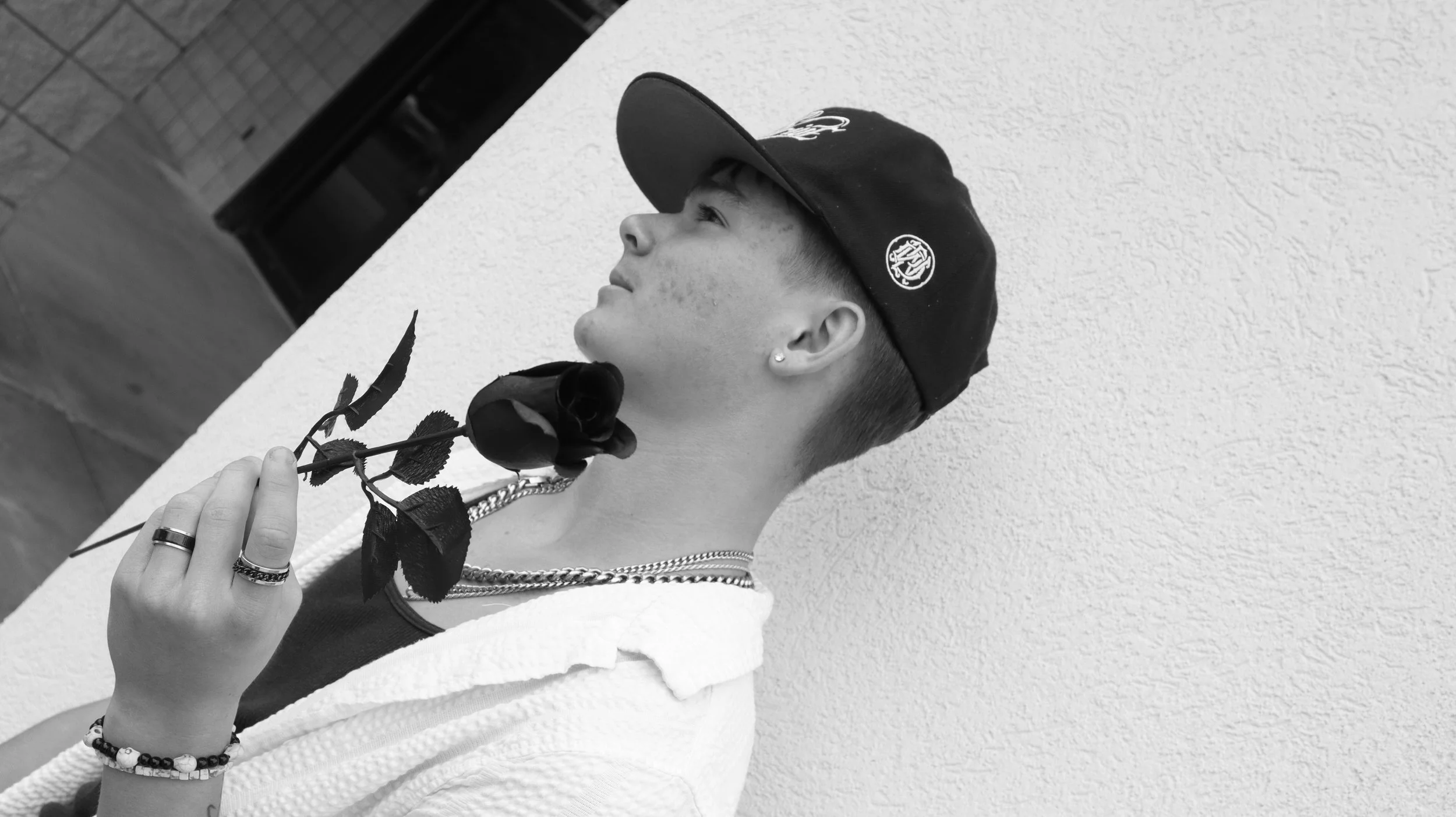 A young person in profile holding a black rose, wearing a cap, jewelry, and a white garment, standing against a textured wall.