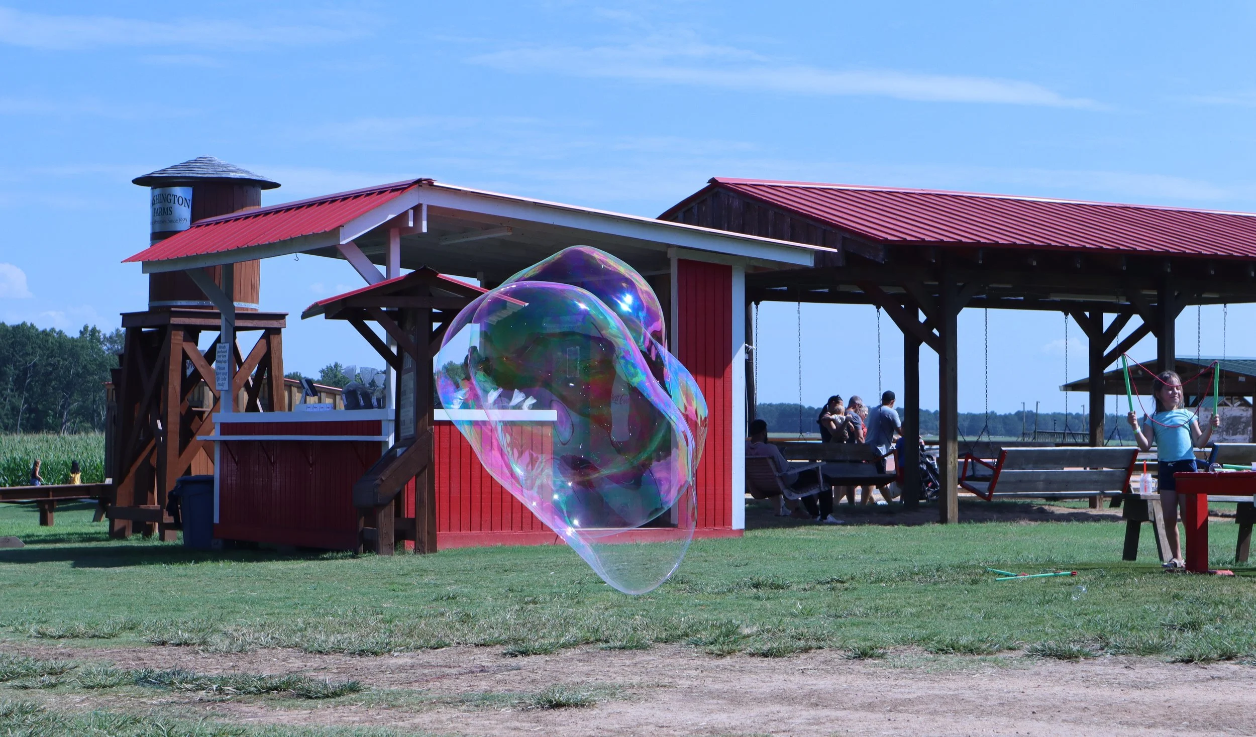 A park with a red and white pavilion, children, adults, and a large soap bubble in the foreground, green grass, trees, and a blue sky in the background.