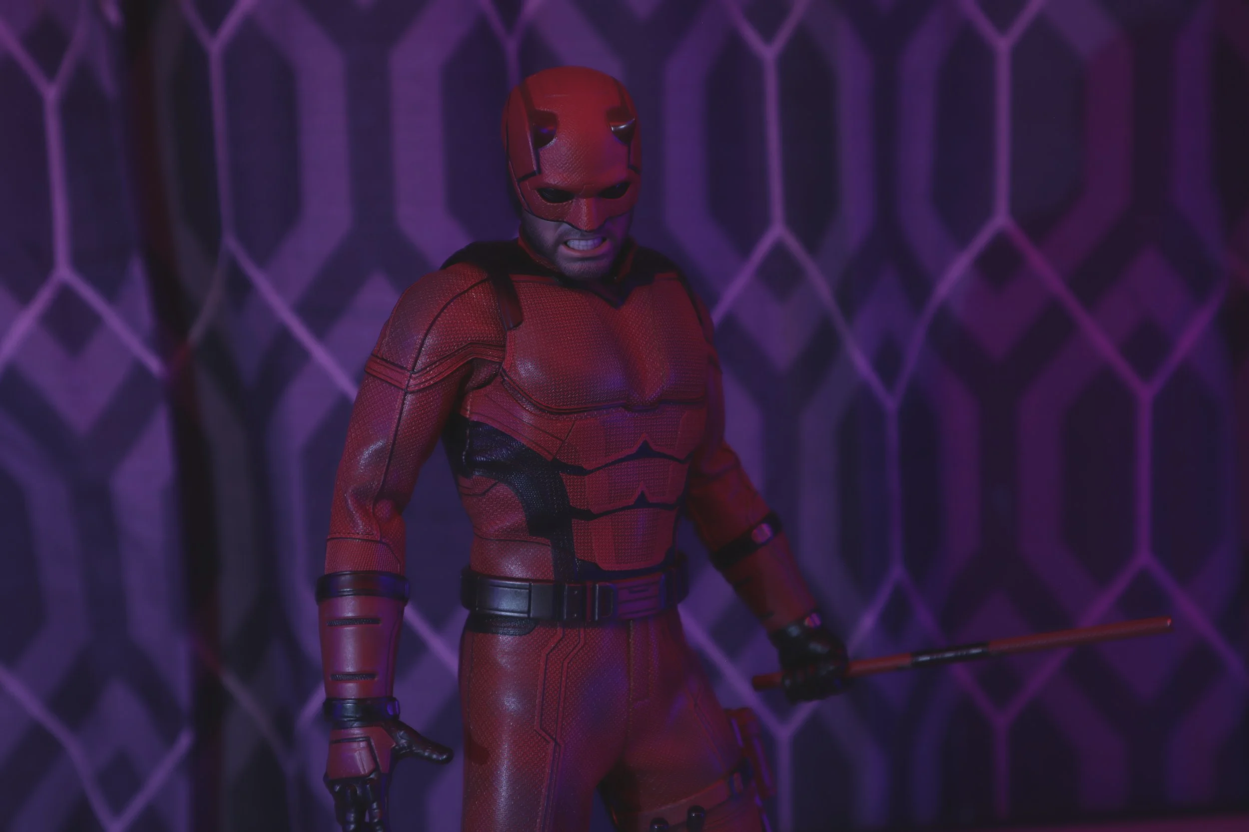 Hot Toys Daredevil in a red and black costume holding a weapon, standing in front of a purple patterned background.