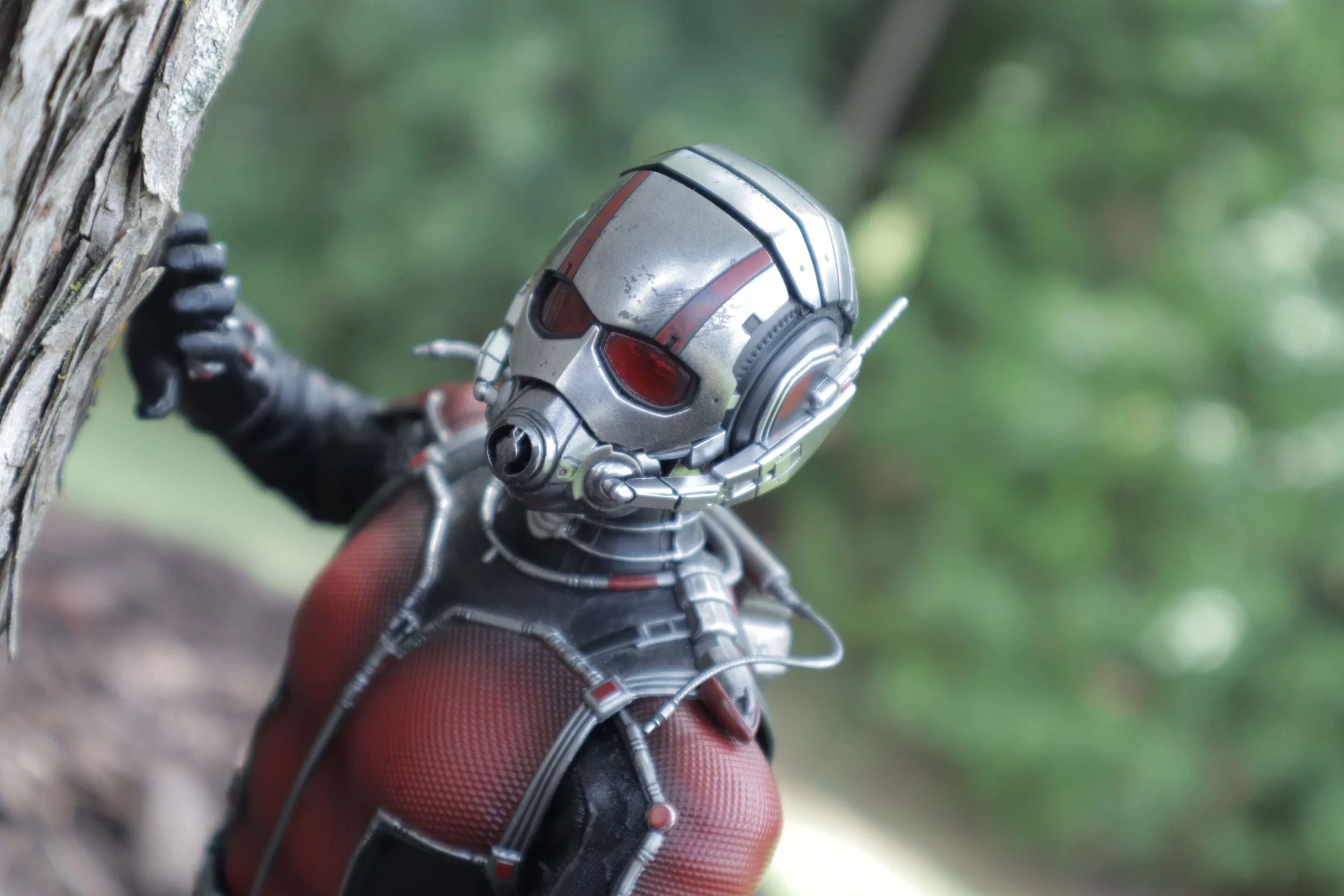 Close-up of a silver and red futuristic robot or superhero costume with a helmet, gloves, and arm armor, reaching out and touching a tree trunk outdoors with a blurred green background.