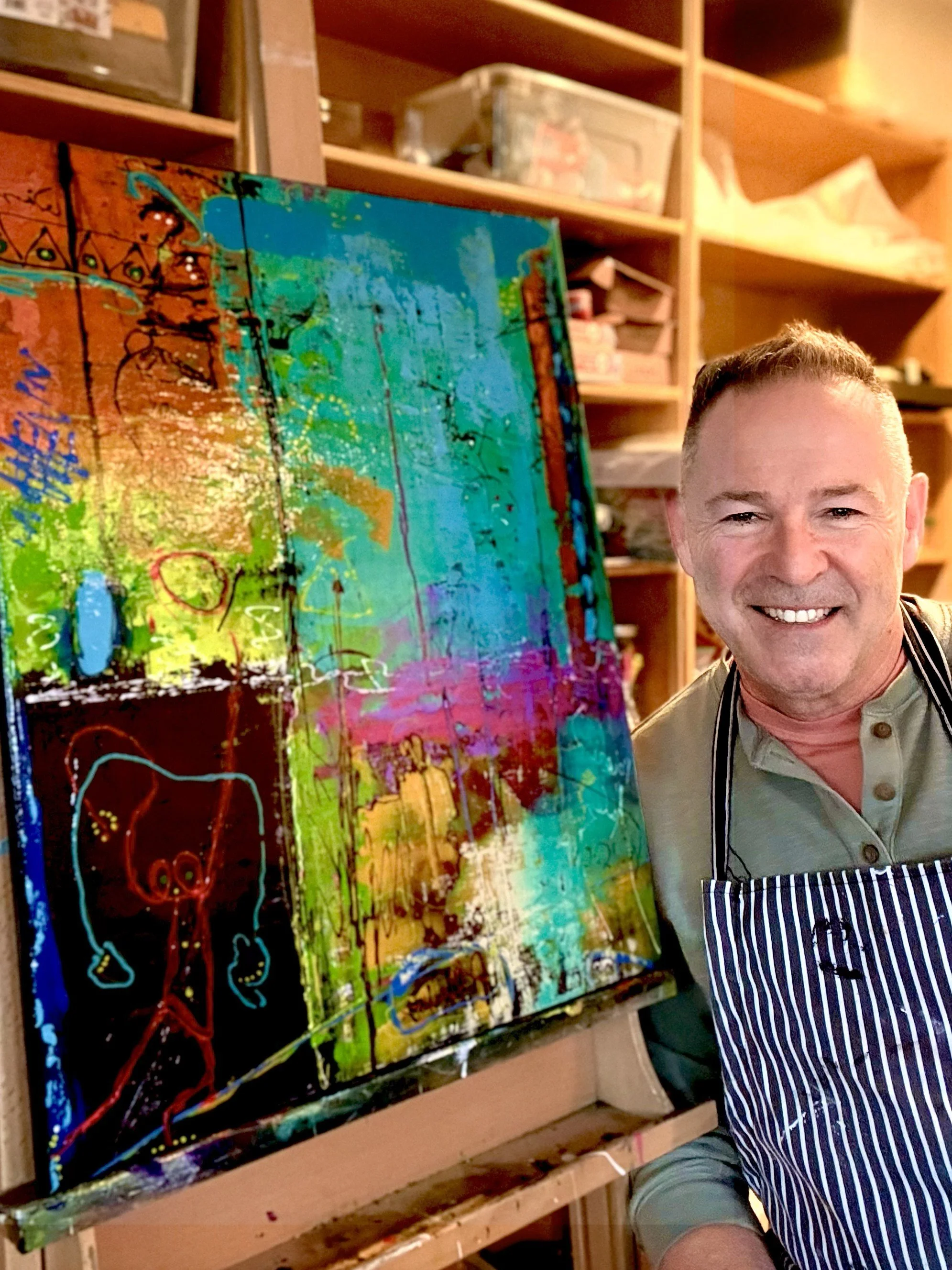 Artist Aedan Hanley standing beside a colorful mixed-media painting in his studio.