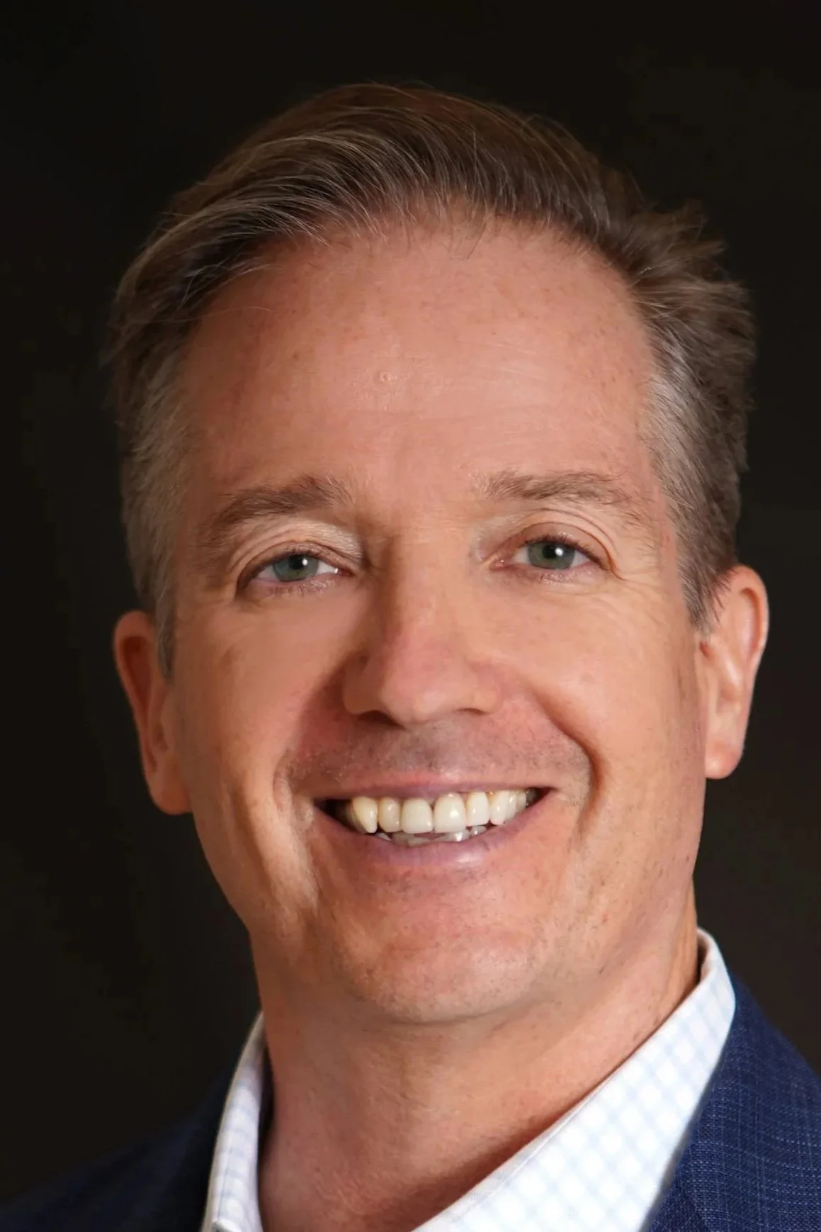 Close-up of smiling middle-aged man with short brown hair, blue eyes, wearing a navy blazer and light blue checkered shirt against a dark background.