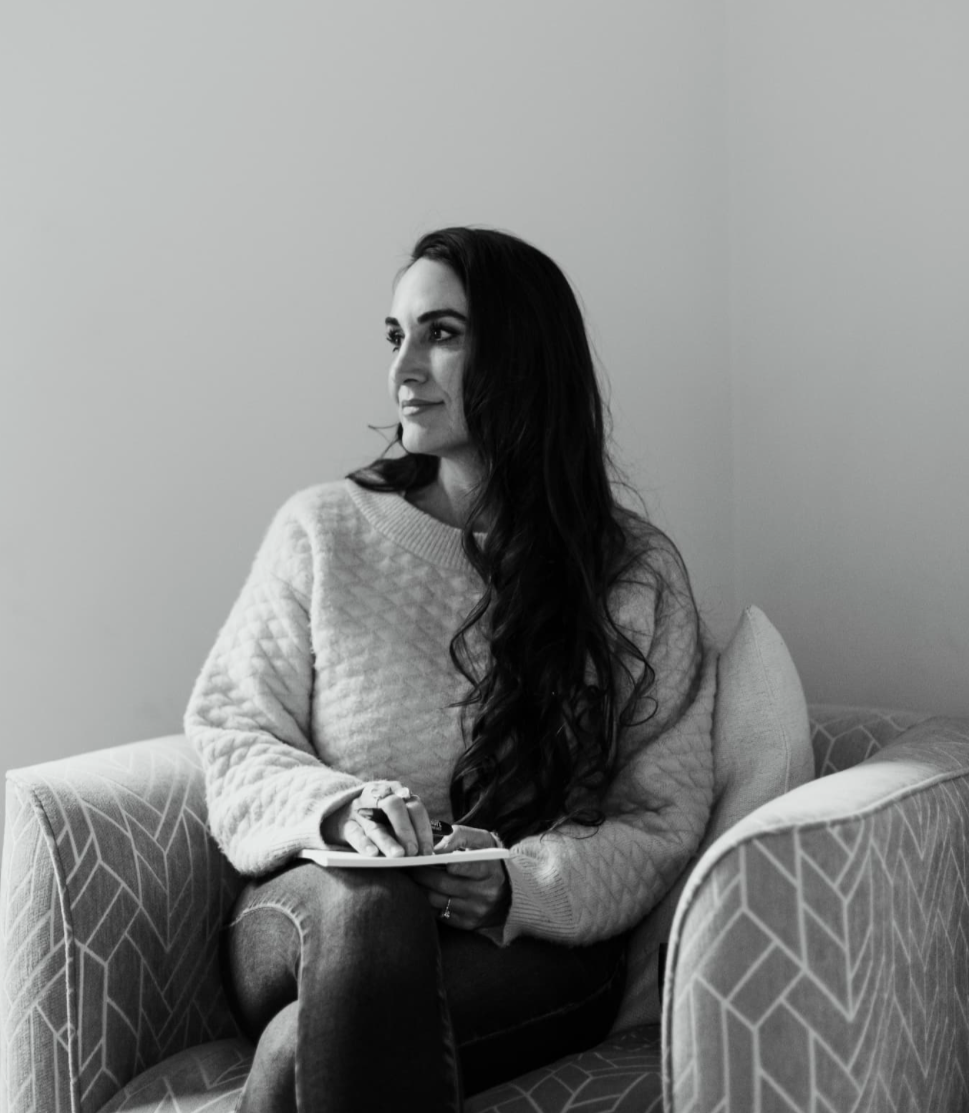 A woman with long dark hair sitting on a patterned armchair, holding a notepad and pen, looking to the side in a room with plain walls.