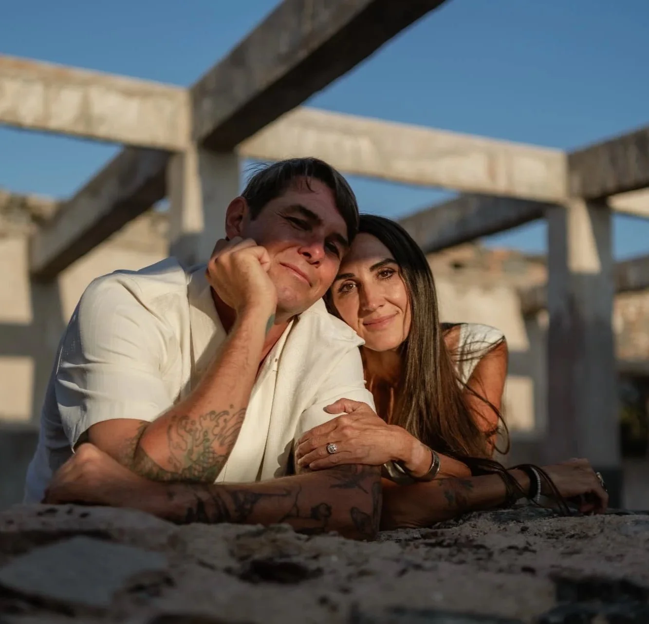A man with tattoos resting his chin on his hand and a woman with long dark hair smiling, both laying on a beach with wooden structures and clear blue sky in the background.