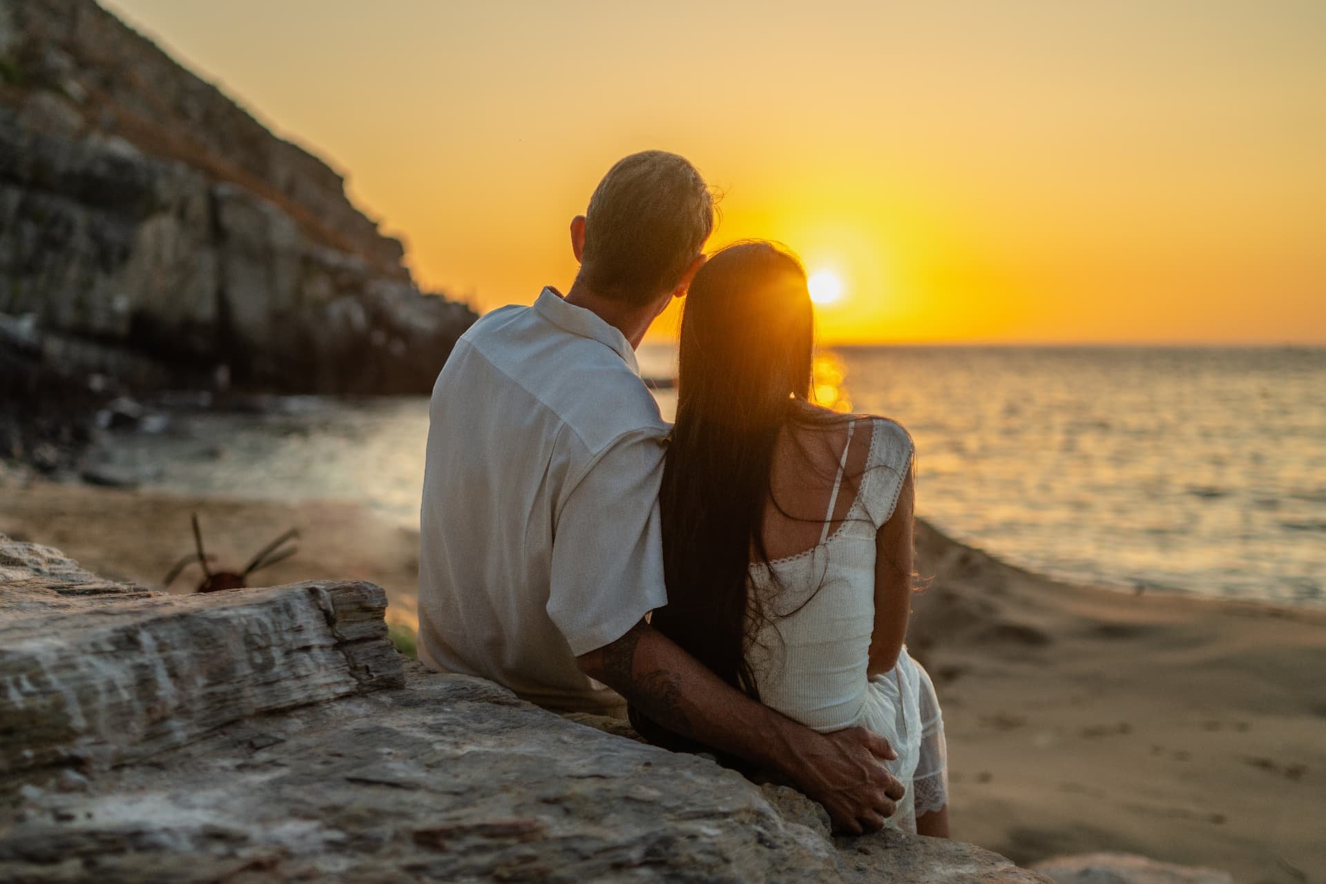 A couple sitting on a beach during sunset, looking at the horizon, with the ocean in front and a rocky cliff on the left.