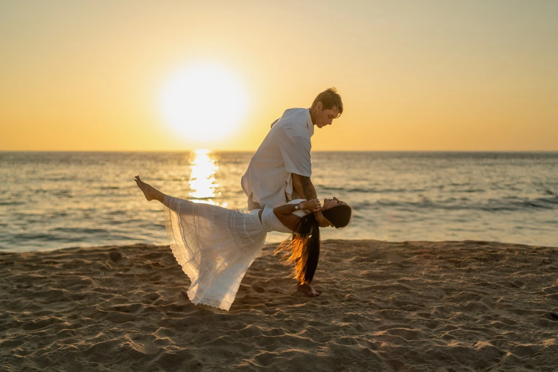 A couple dancing on the beach during sunset, with the man holding the woman in a dip. The sun is low on the horizon, reflecting on the water, creating a warm, golden glow.