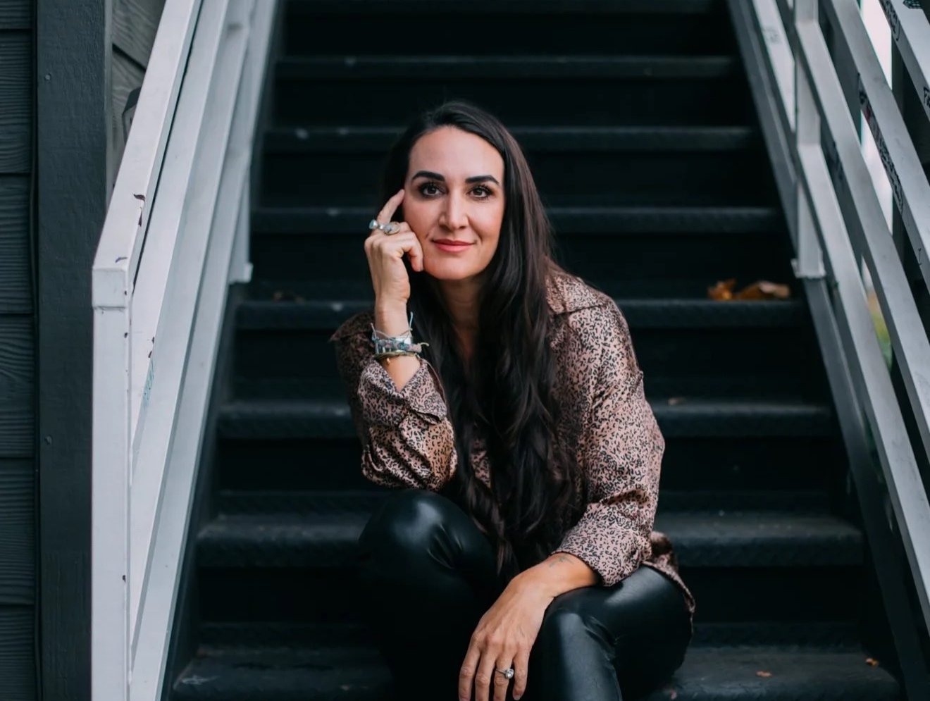 A woman sitting on outdoor stairs, resting her chin on her hand, wearing a leopard print shirt and black leather pants.