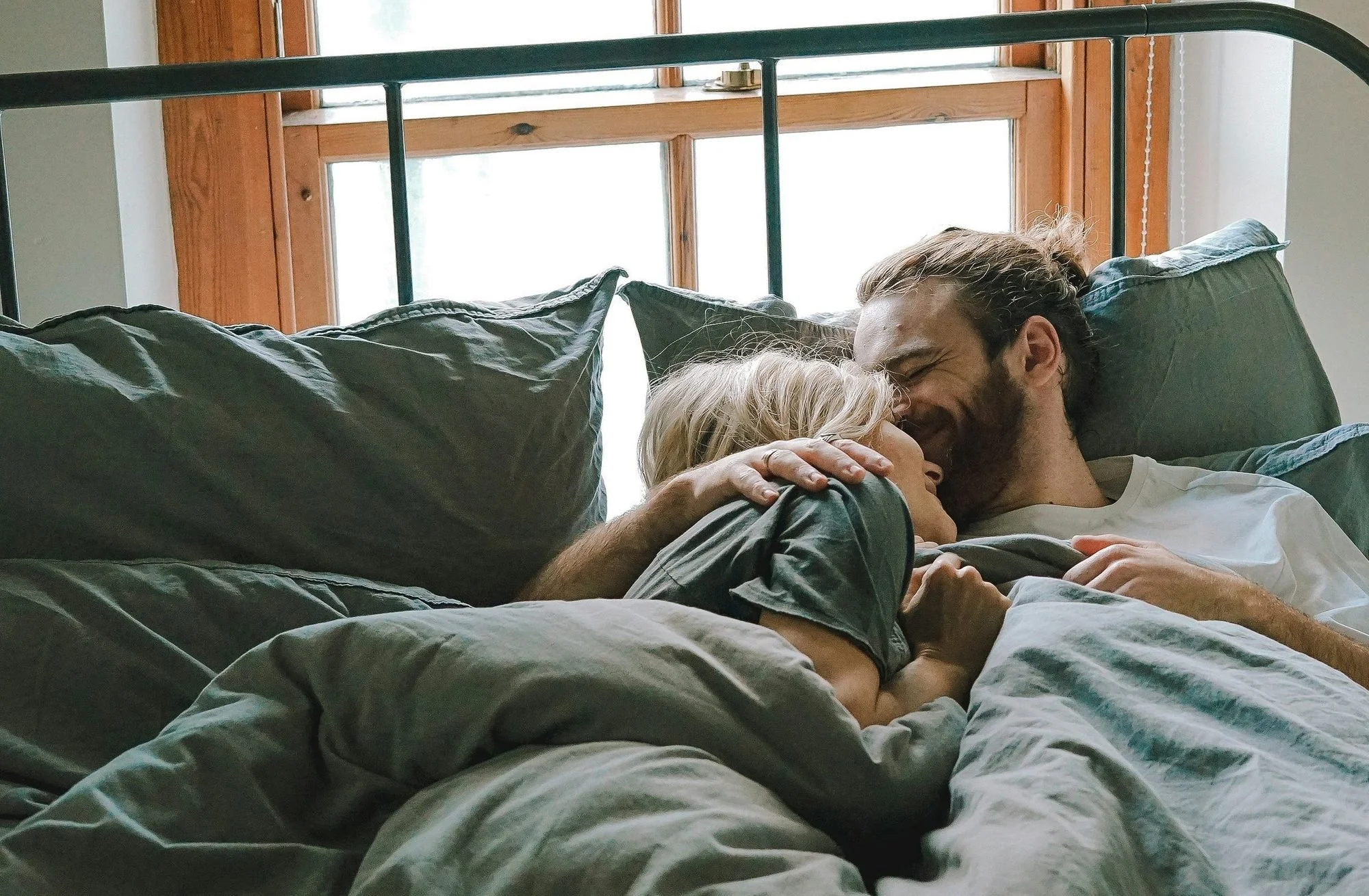 A man and a woman lying in a bed, smiling, and about to kiss in a cozy bedroom with a wooden window behind them.