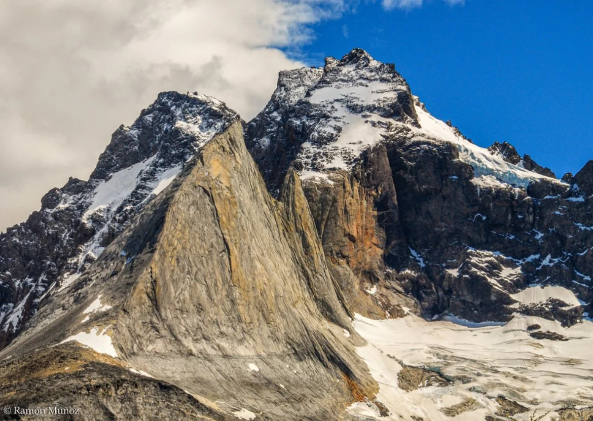 A rugged mountain landscape with snow-capped peaks, rocky slopes, and patches of ice, under a partly cloudy sky.