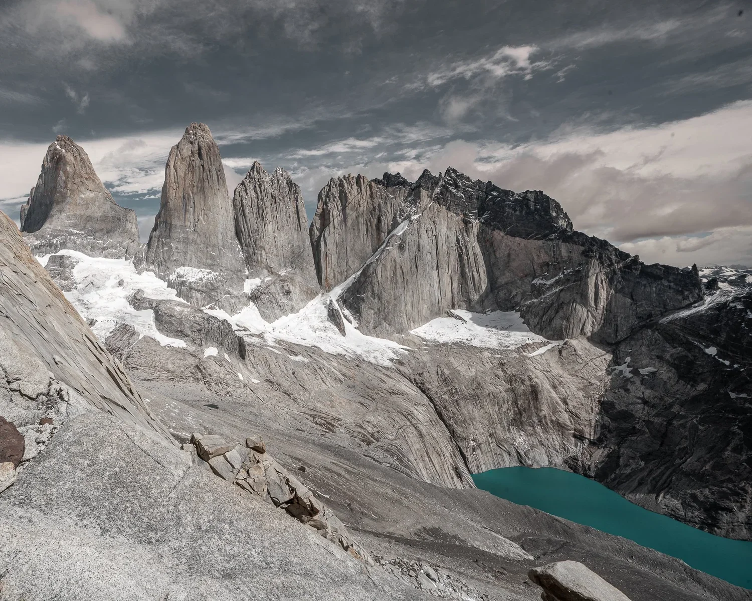 A rugged mountain landscape with tall, jagged peaks, snow patches, and a small turquoise lake at the base, under a cloudy sky.
