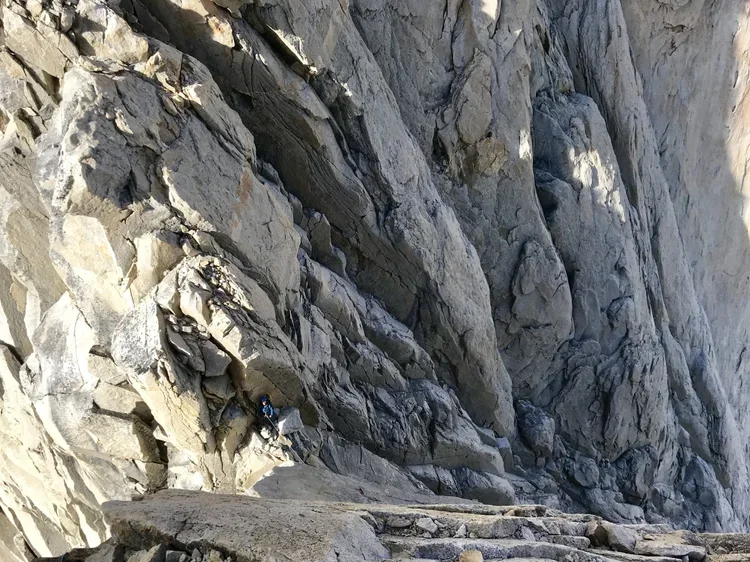 Climber in blue gear ascending a rocky cliffside with steep, jagged gray rocks.