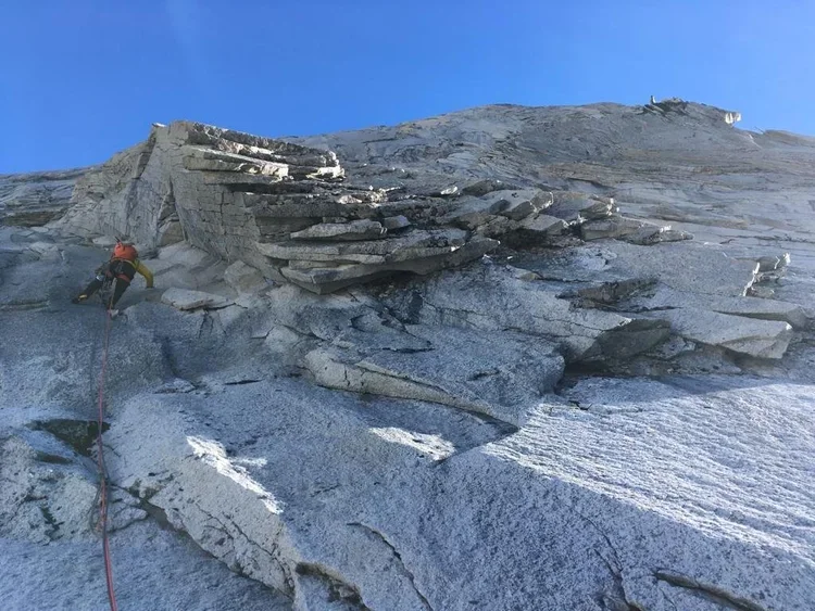 A climber wearing a helmet and harness ascending a rocky and snowy mountain slope with a blue sky background.