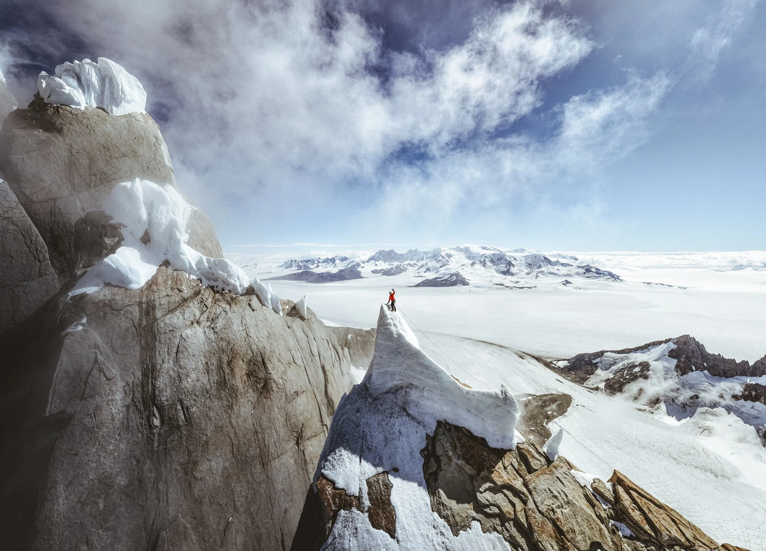 A snow-covered mountain range with a person climbing on rocks in the foreground, under a partly cloudy sky.