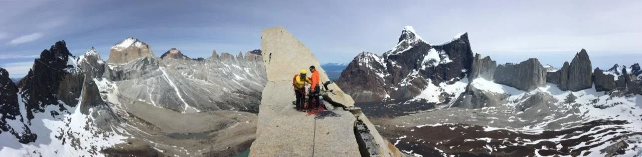 Two climbers sitting on a narrow rocky ledge at a mountain summit with snow-capped peaks around them.