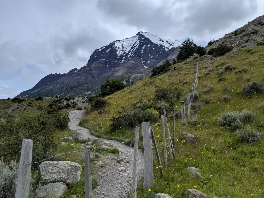 A mountain landscape with a winding dirt trail, a fence along the trail, grassy hills with shrubs, and a snow-capped mountain in the background under cloudy skies.
