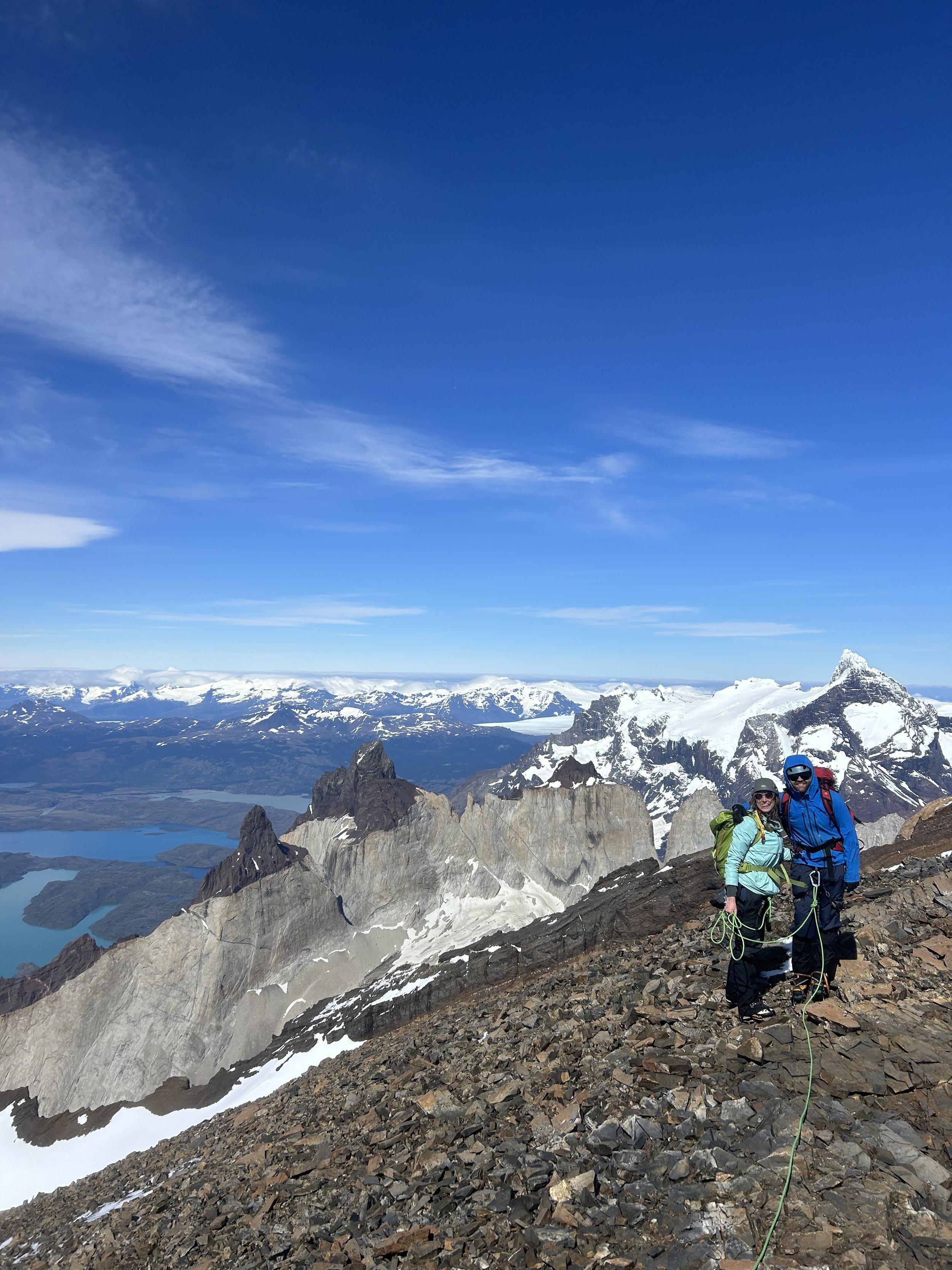 Two hikers in colorful gear standing on rocky mountain terrain with snow-capped peaks and a blue sky in the background.
