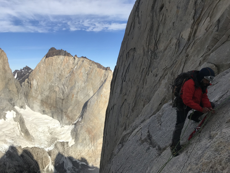A climber in red jacket and helmet ascending a steep granite cliff with mountains and partly cloudy sky in the background.