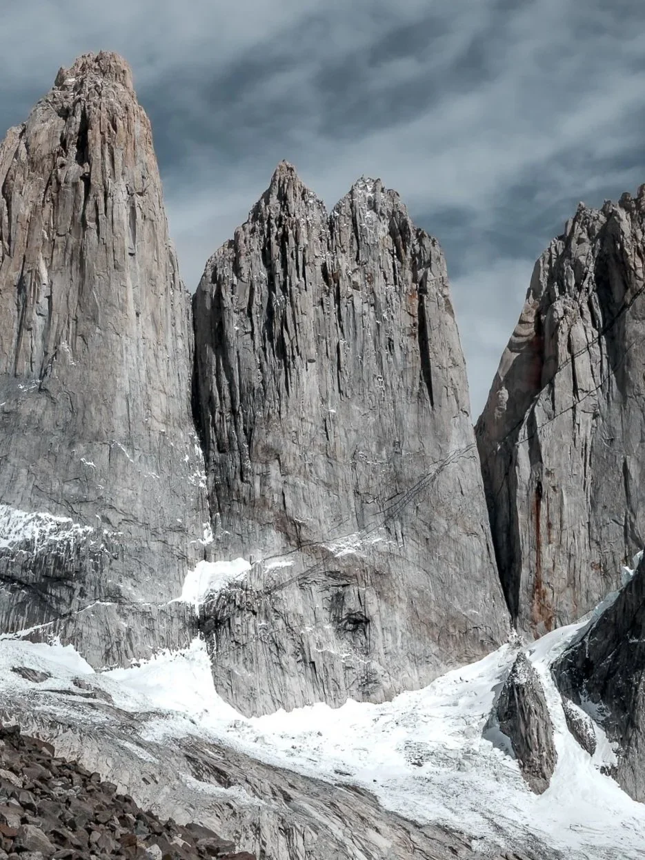 Tall, jagged rocky mountain peaks with snow and ice at the base under a cloudy sky.