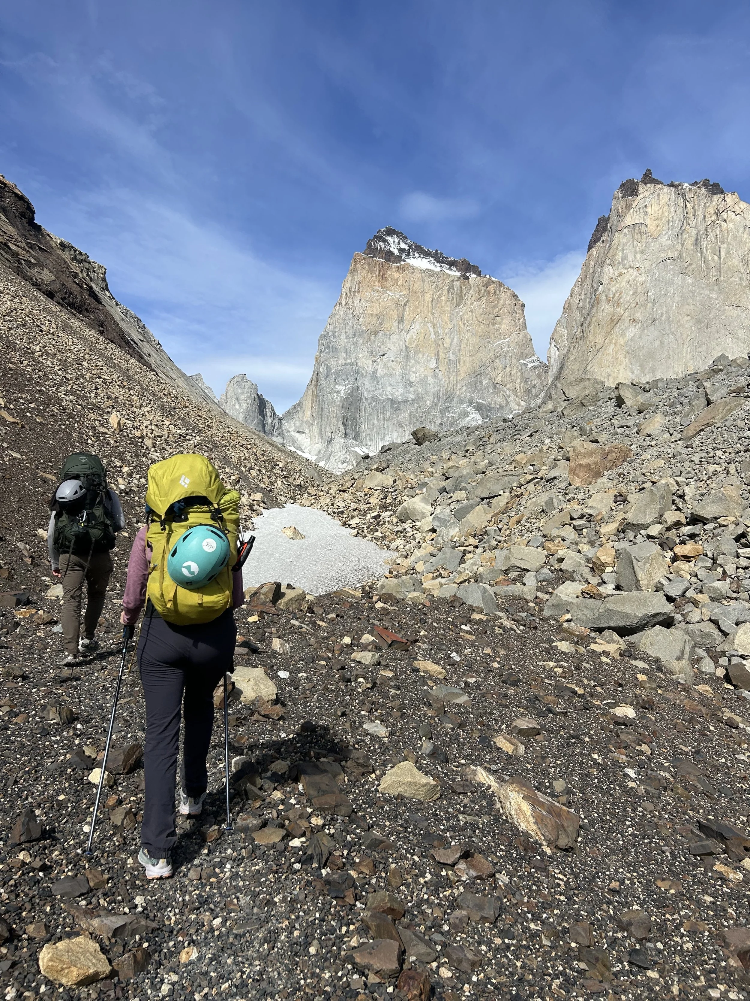 Group of hikers walking on rocky trail in mountainous area with tall cliffs and snow patches, under blue sky.