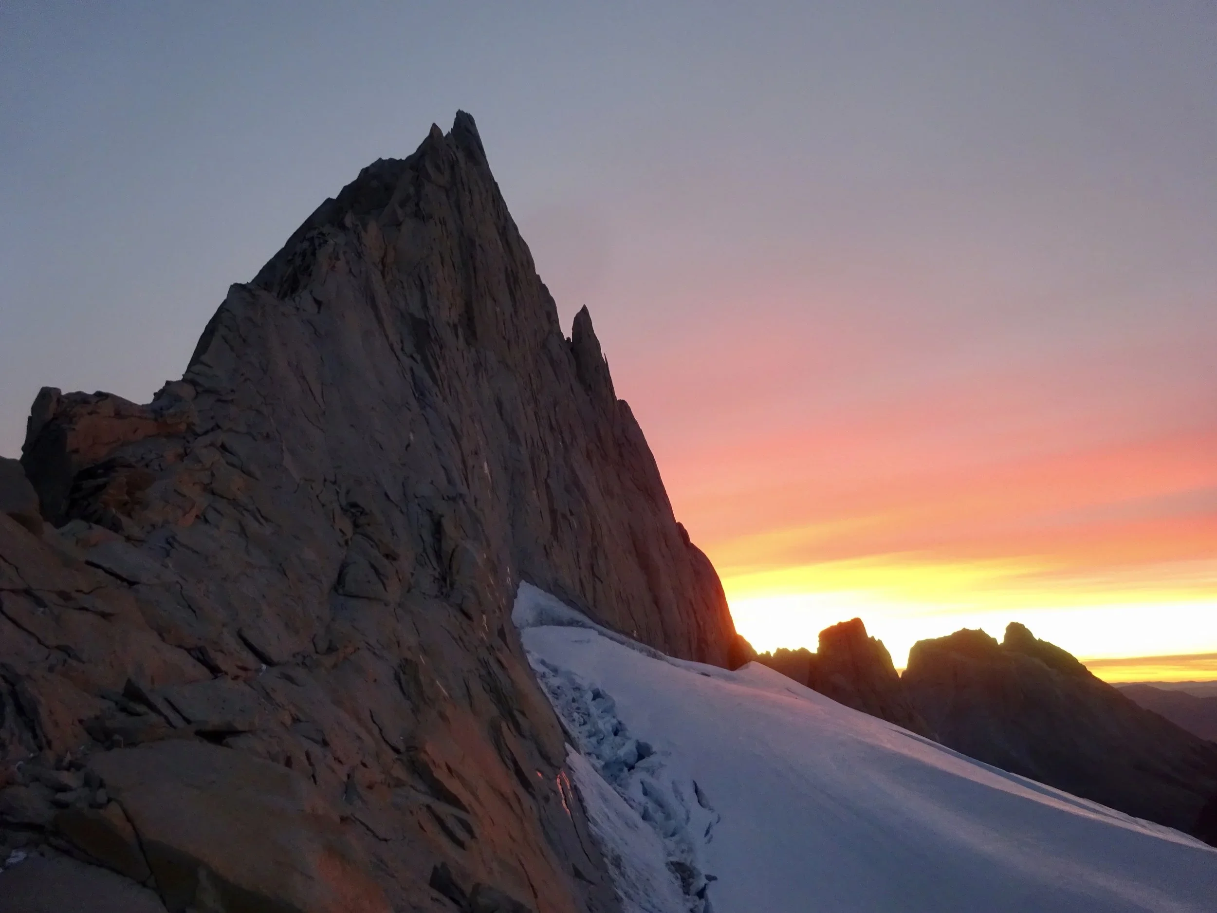 Snowy mountain slope at sunset with colorful sky and rugged peaks in the distance.