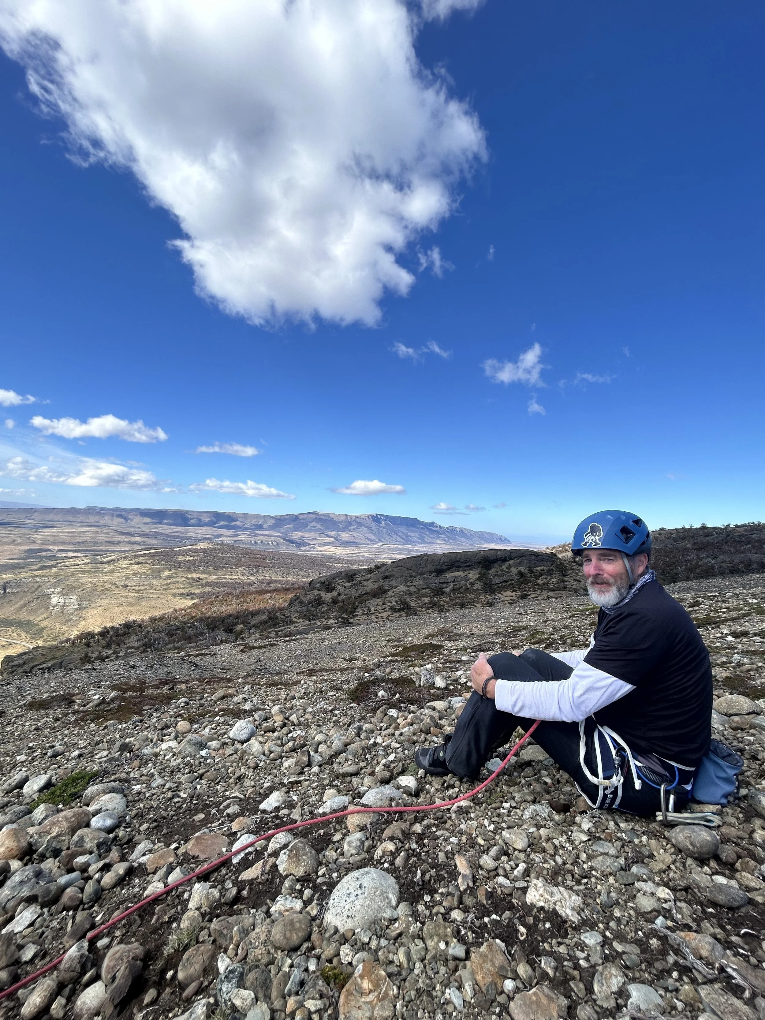 A man with a beard wearing a blue bicycle helmet and sitting on a rocky hillside, with a mountain range and blue sky with scattered clouds in the background.