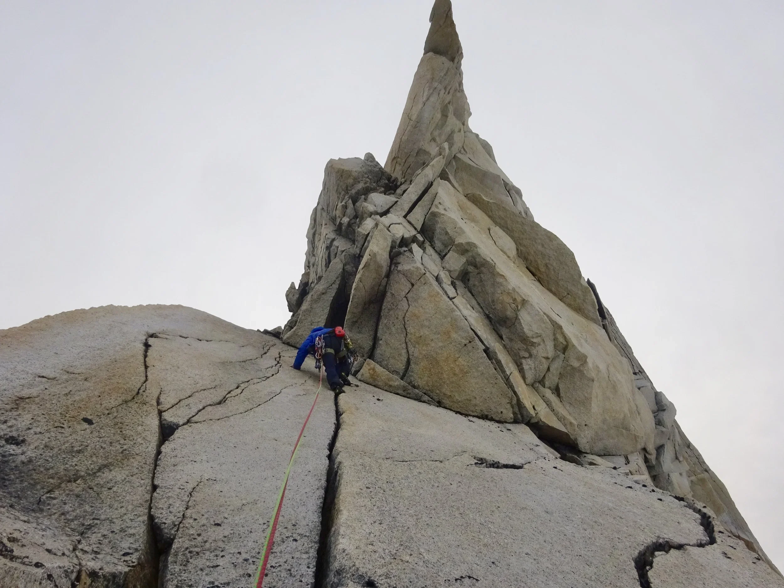 Climber with helmet and harness ascending a steep, cracked granite rock face toward a jagged peak, with an overcast sky in the background.
