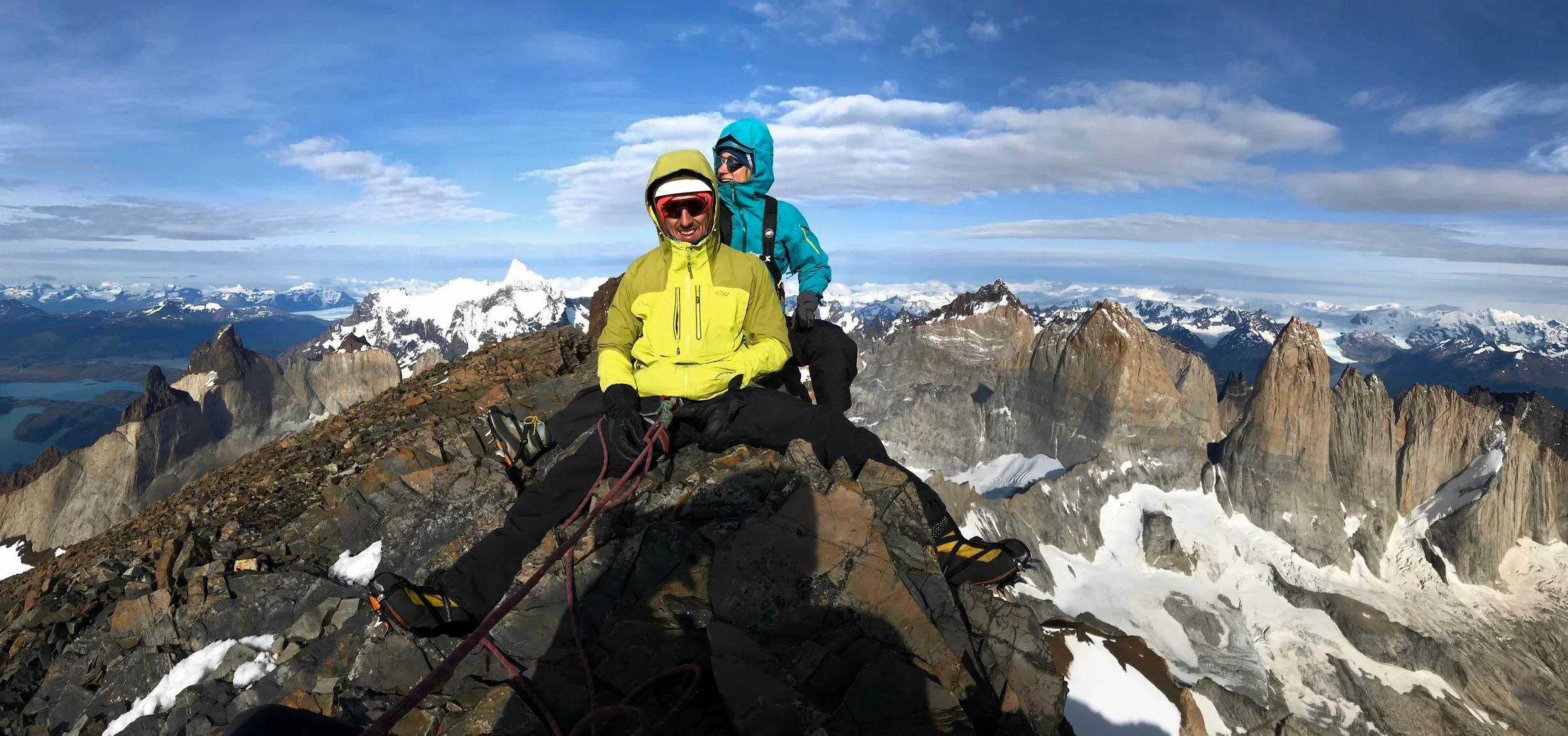 Two climbers resting on a rocky mountain peak surrounded by snow-capped peaks and a blue sky with clouds.