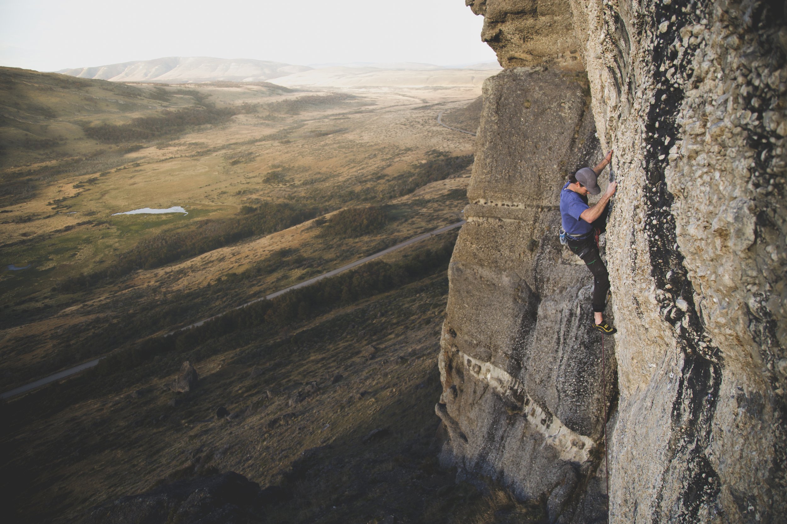 A person rock climbing on a steep vertical cliff face in a remote mountainous area, with open land and hills in the background.