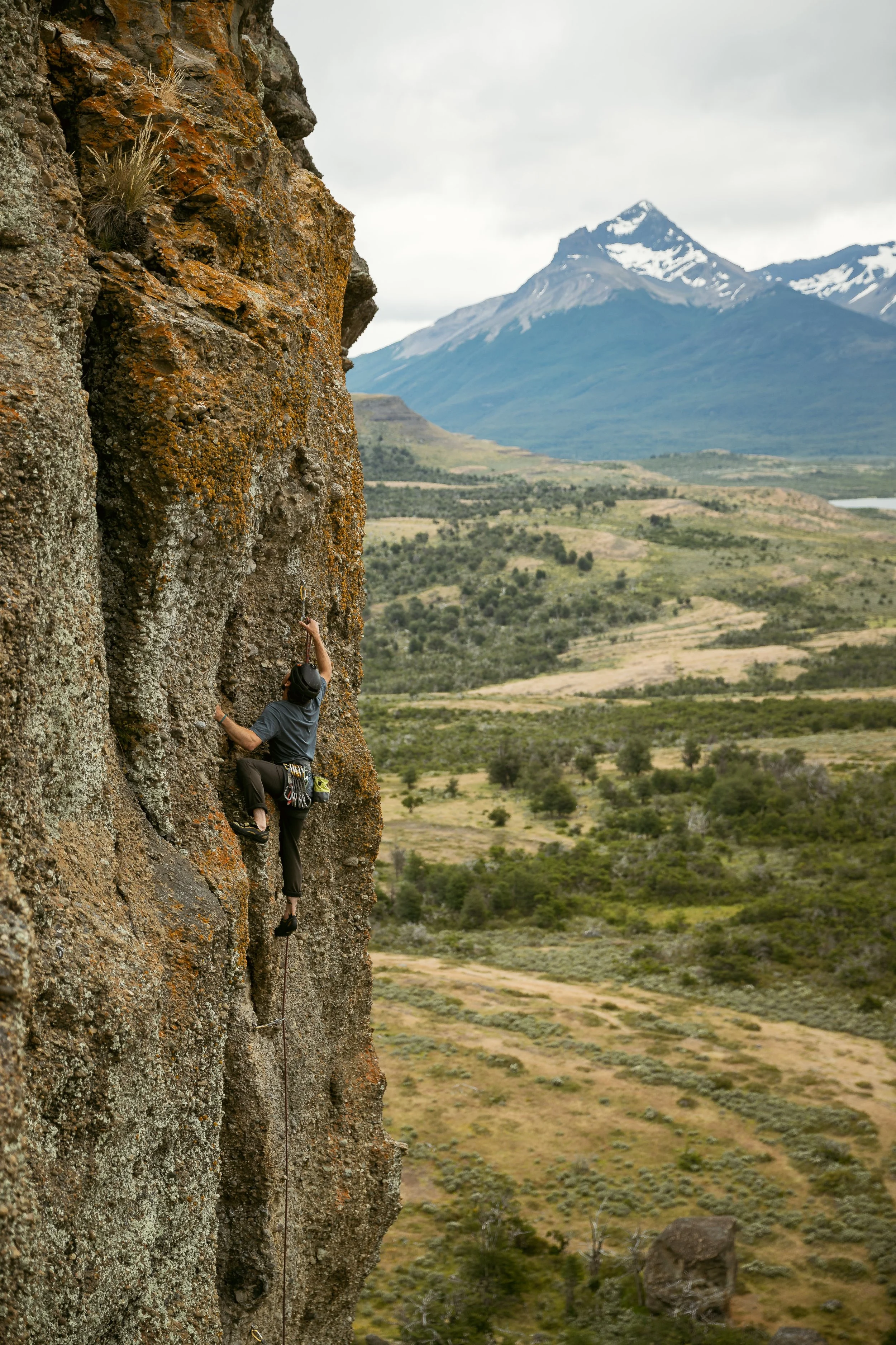A person rock climbing on a rugged cliff with a mountain range in the background.