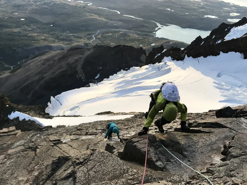 Two climbers ascending a snowy mountain ridge with a scenic landscape and lakes in the background.
