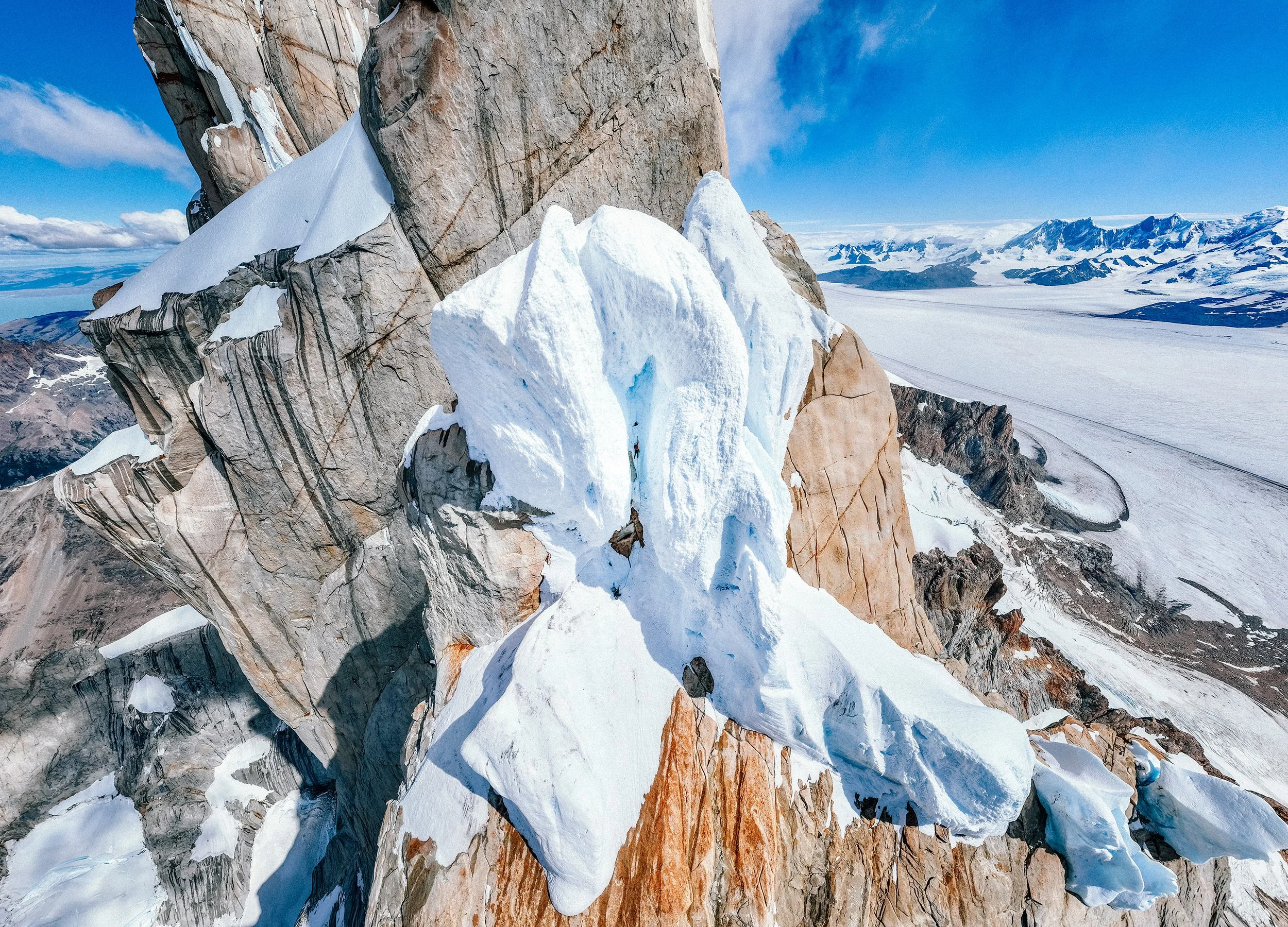 Snow-covered mountain summit with rocky cliffs against a blue sky and distant mountain ranges.