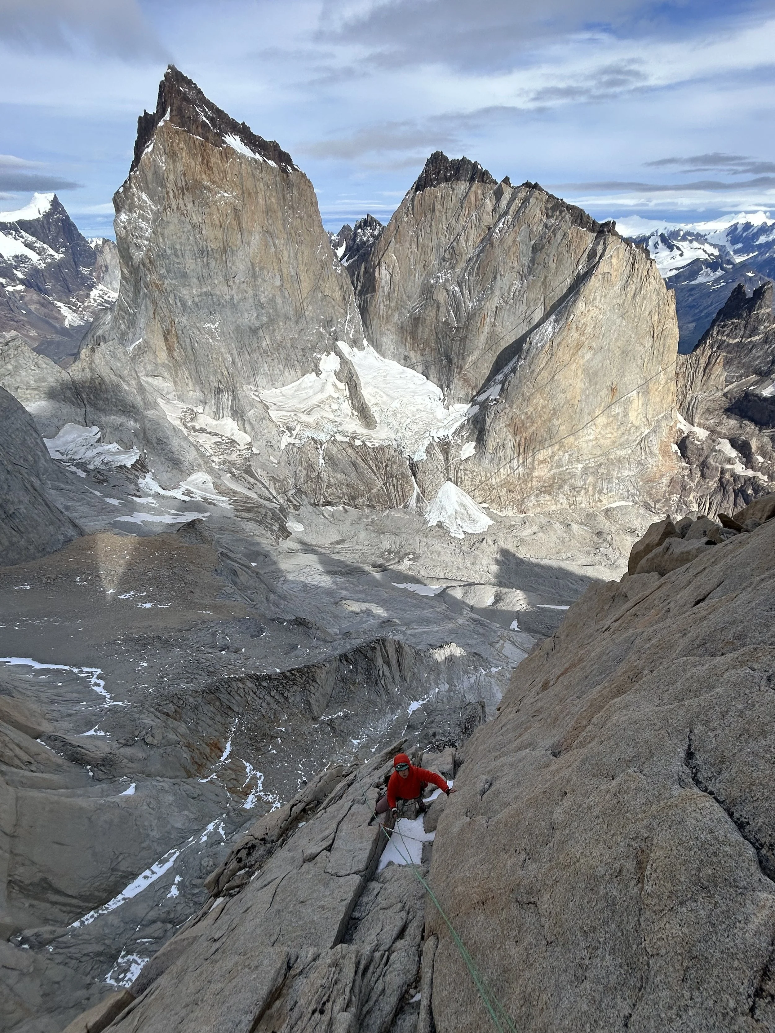 A person climbing a steep granite rock face in a mountainous area with snow and jagged peaks in the background.