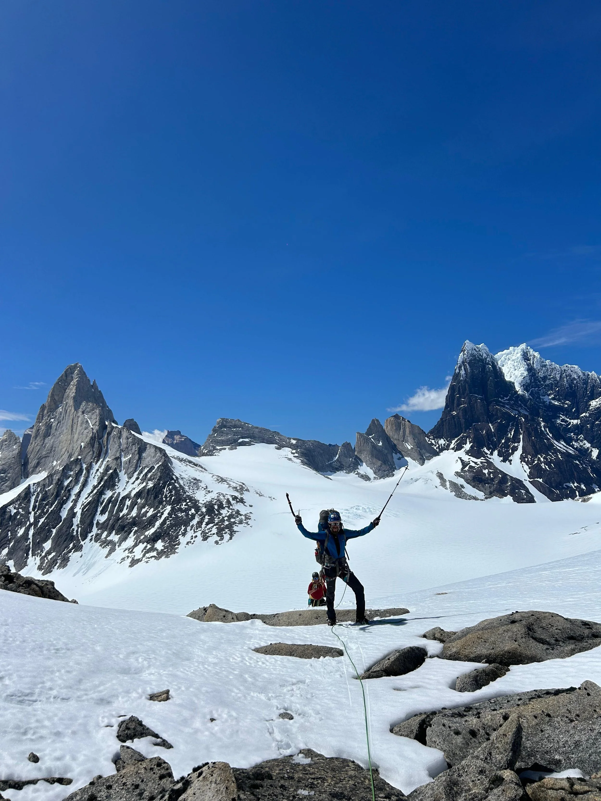 A person dressed in winter gear, wearing a helmet and sunglasses, standing on snow with arms raised, holding ski poles, in a mountainous snowy landscape with rocky peaks and a clear blue sky.