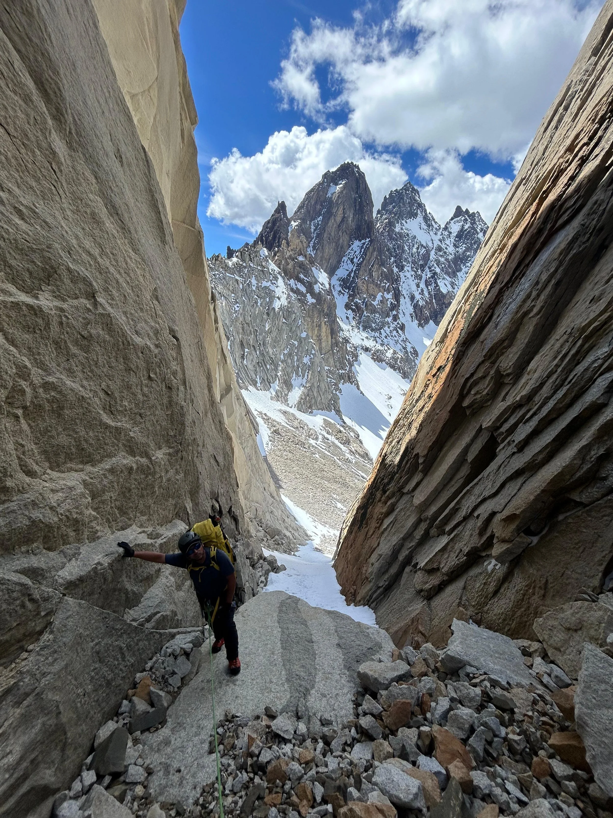 A person in climbing gear and a helmet ascending a steep, narrow mountain gorge filled with snow and rocks, with towering snow-capped mountain peaks in the background against a blue sky with scattered clouds.