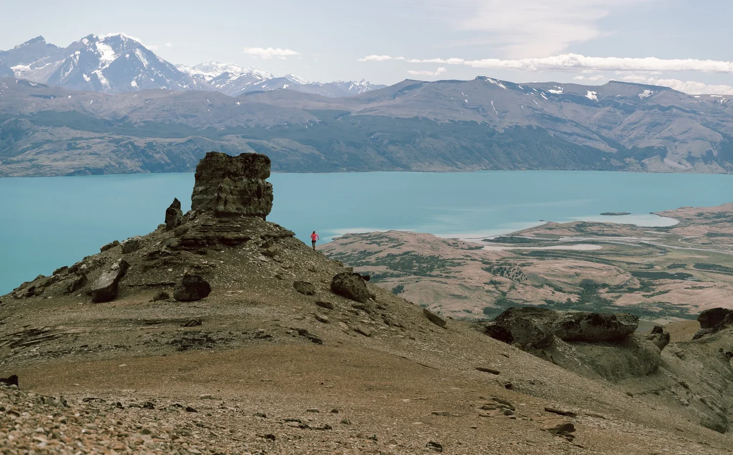 A person standing near a rock formation on a hill overlooking a large lake with mountains in the background.