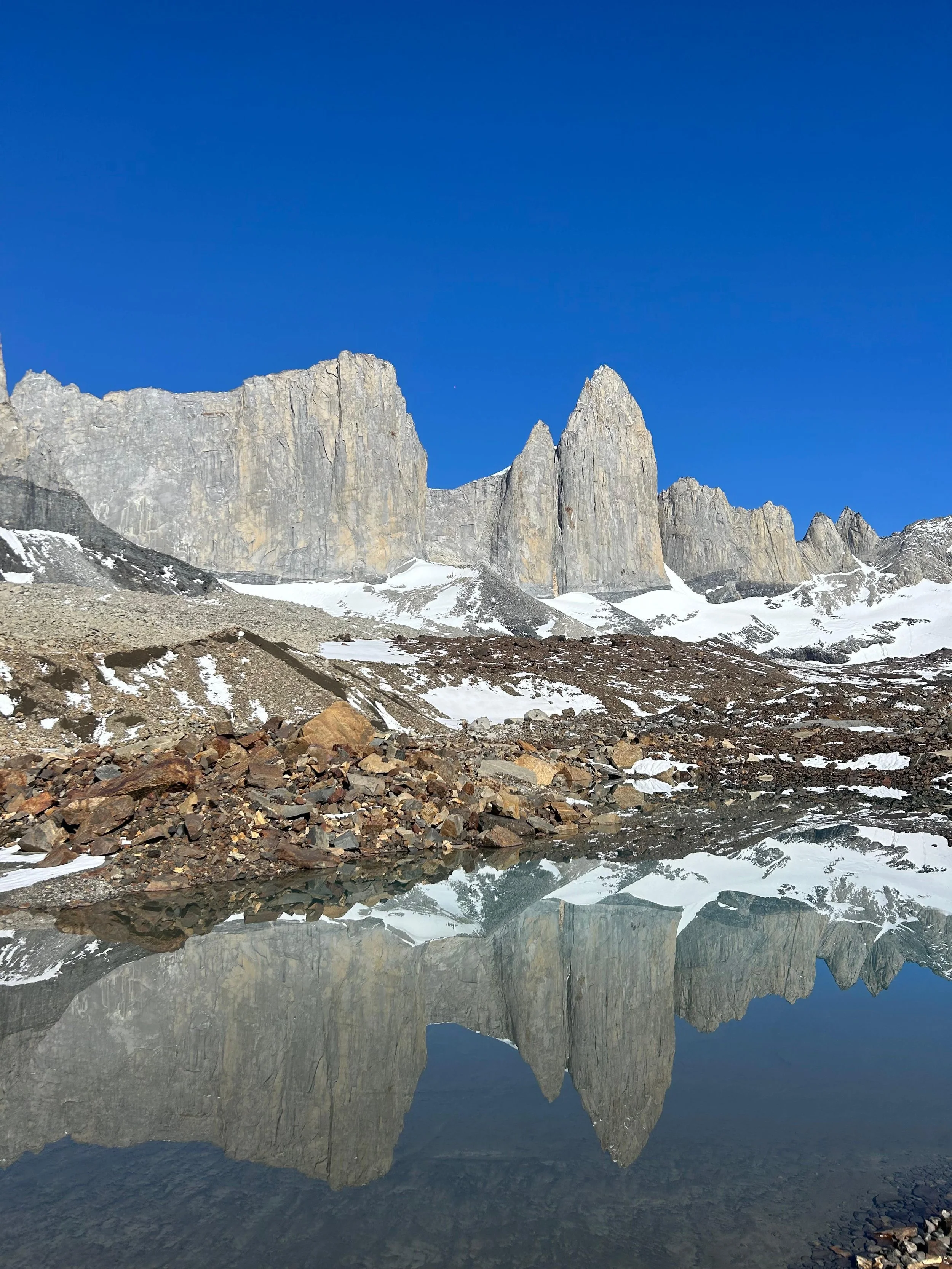 Snow-capped mountain peaks with rocky foreground and clear blue sky, reflected in a calm body of water.